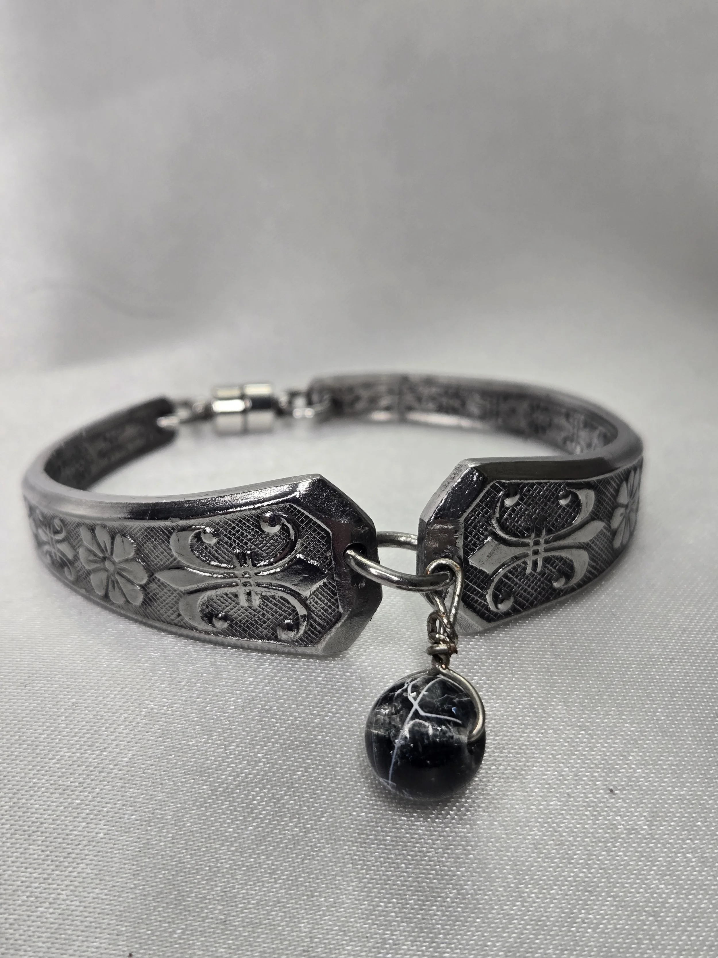 A black and silver bracelet with floral patterns and a dangling black and silver pendant, set on a light gray textured surface.