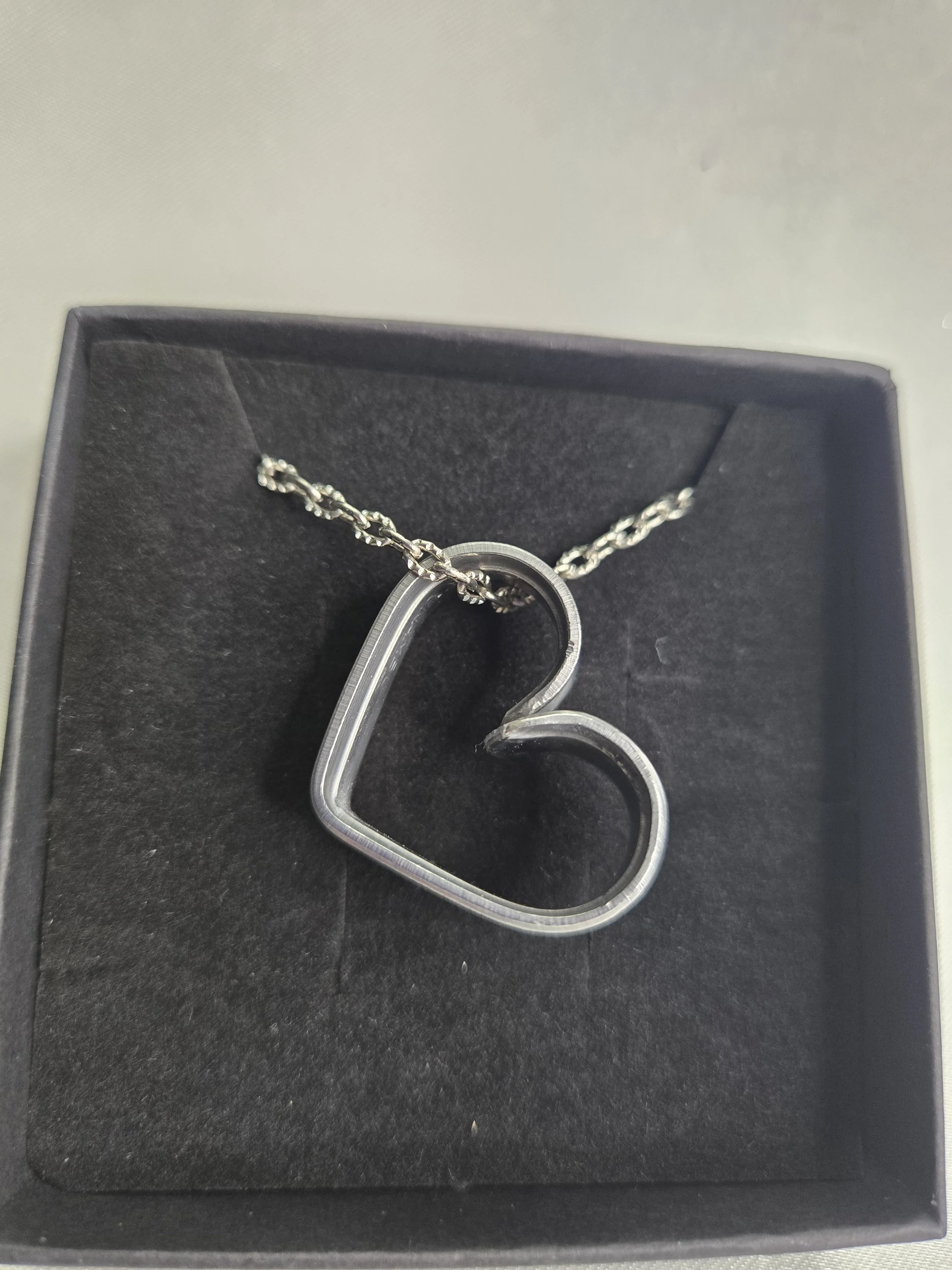 A silver heart-shaped necklace with a chain displayed on a black velvet surface inside a black jewelry box.