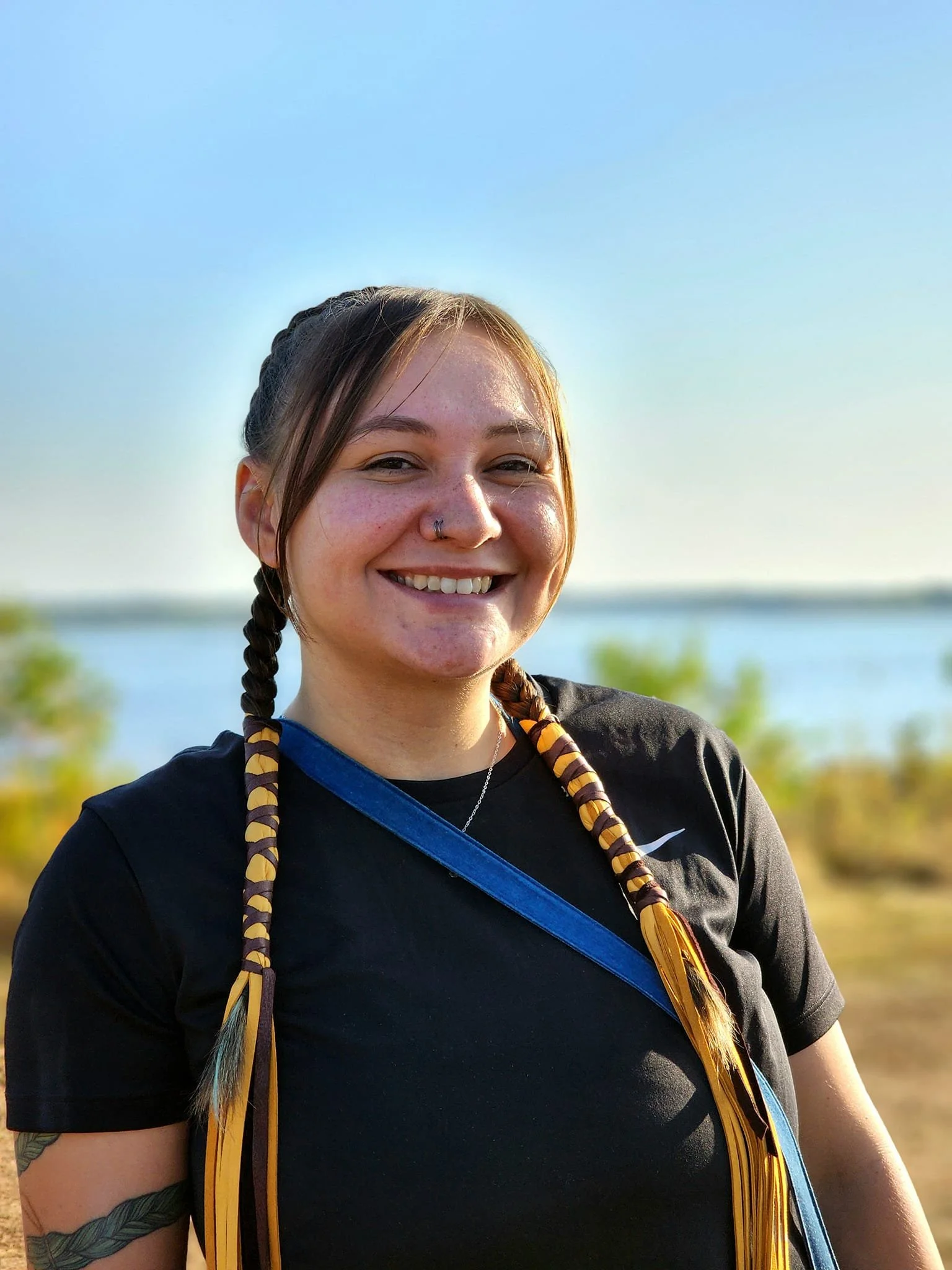 A young woman with braided hair, wearing a black Nike shirt and smiling outdoors near a body of water and green trees.