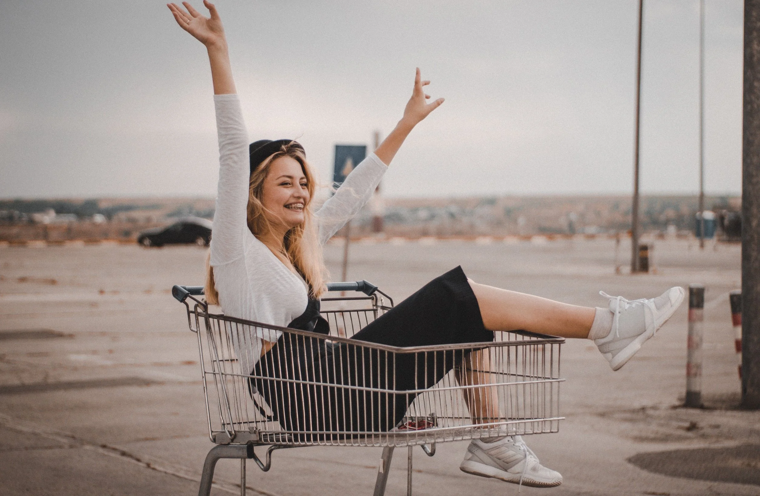 A young woman with blonde hair smiling and raising her arms while sitting in a shopping cart outdoors, with a parking lot and distant cityscape in the background.
