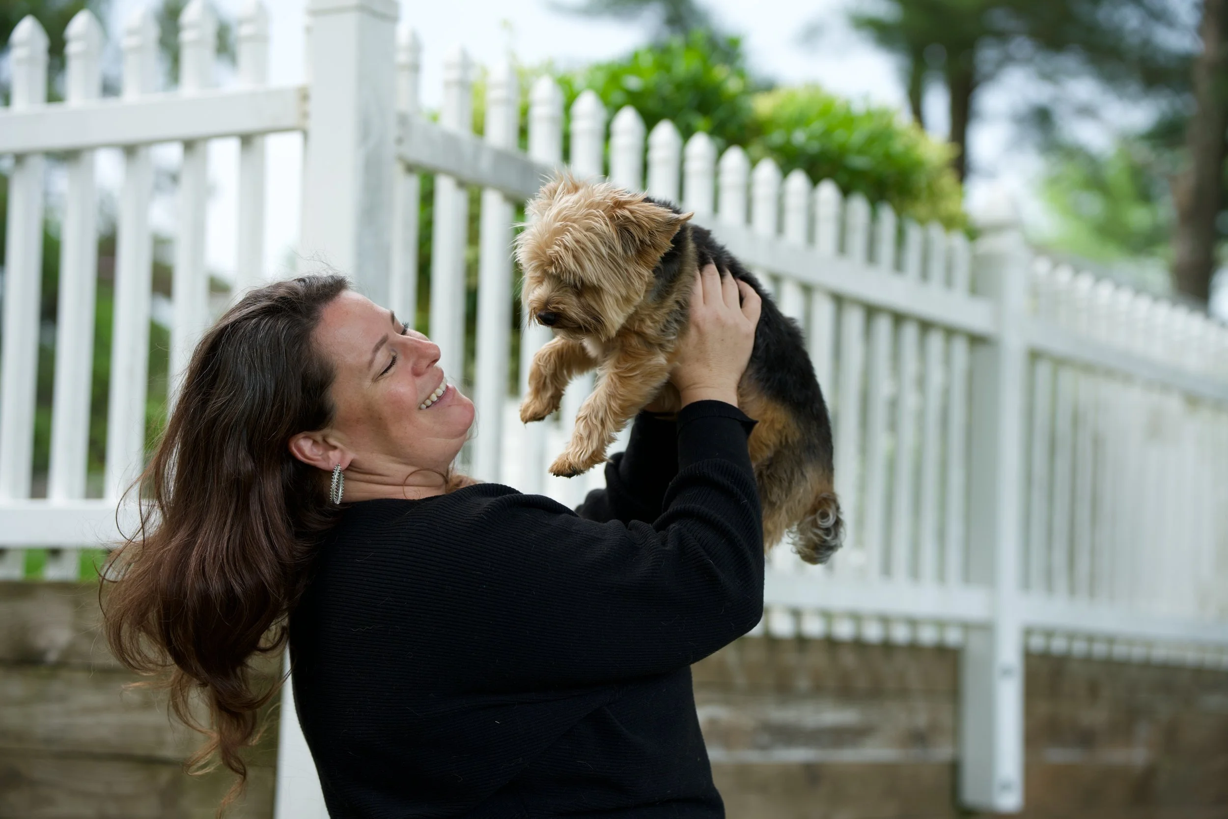 A woman with long brown hair holding a small Yorkshire Terrier puppy up in front of a white picket fence outdoors, smiling at the dog.