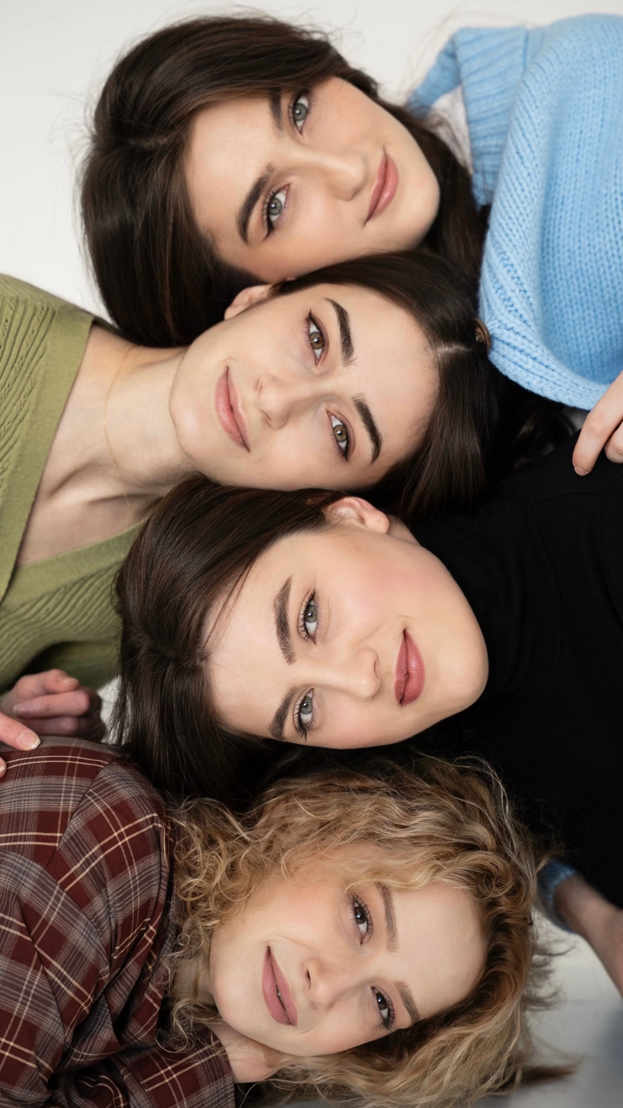 Group of seven women lying closely together on a white surface, faces visible, showcasing diverse hair colors and styles, with a relaxed and happy demeanor.