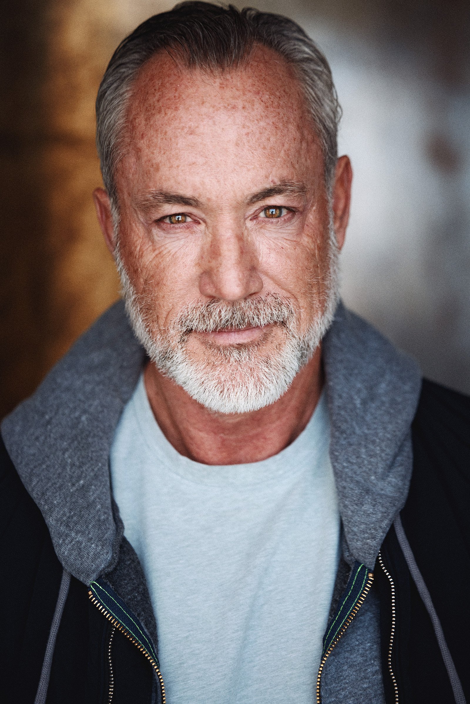 Close-up portrait of a middle-aged man with salt and pepper hair and beard, wearing a light gray t-shirt and a dark gray hoodie, smiling softly at the camera.