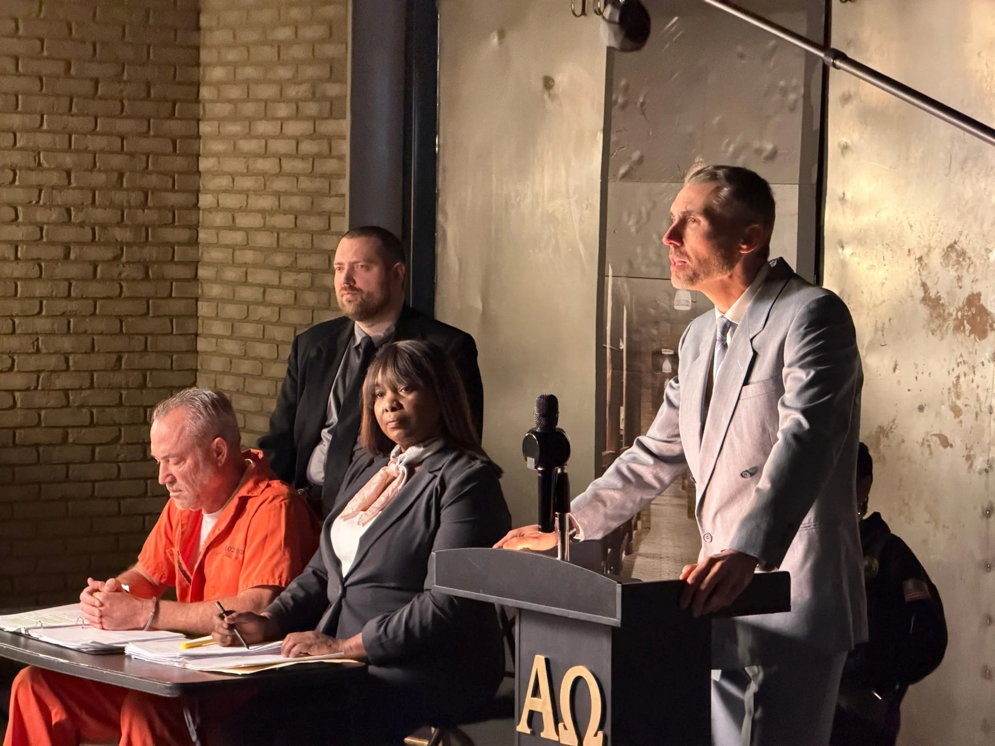 People sitting at a table in a meeting or panel discussion, with one person standing behind a podium, speaking or presenting, in a brick-walled room.