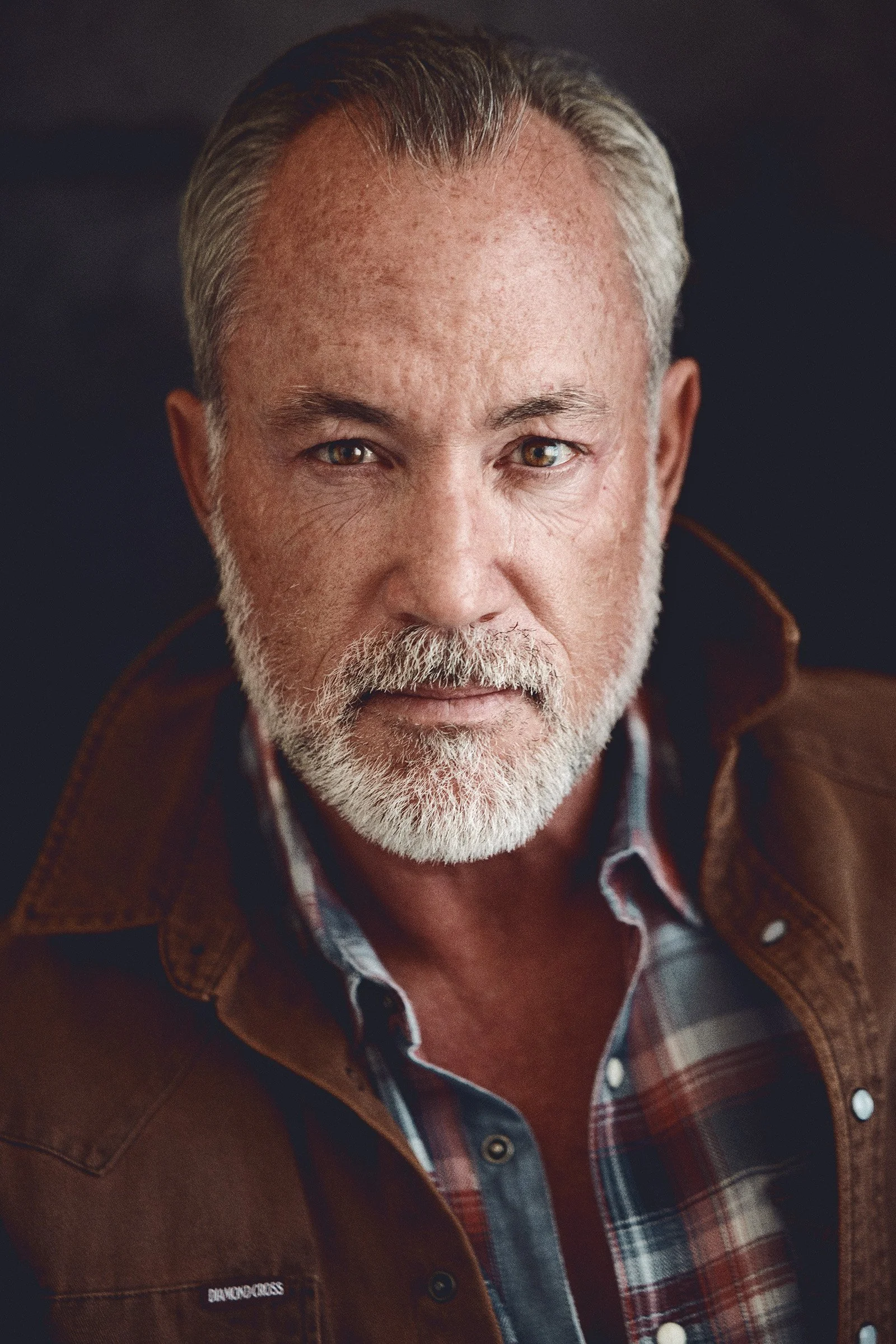Close-up of an man with grey hair and beard, wearing a brown jacket and plaid shirt, looking directly at the camera.