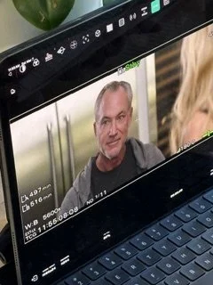 A video call on a laptop screen showing a middle-aged man with short gray hair and a dark shirt, speaking to a woman with blonde hair.