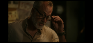 A man with gray hair and glasses adjusting his glasses in a dimly lit room.