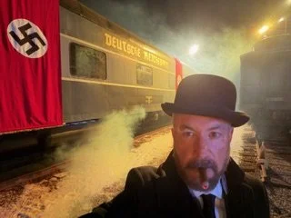 A man wearing a black hat taking a selfie near a train decorated with a Nazi flag and a sign that reads 'Deutsche Bahn.' The train and surroundings are filled with smoke or steam.