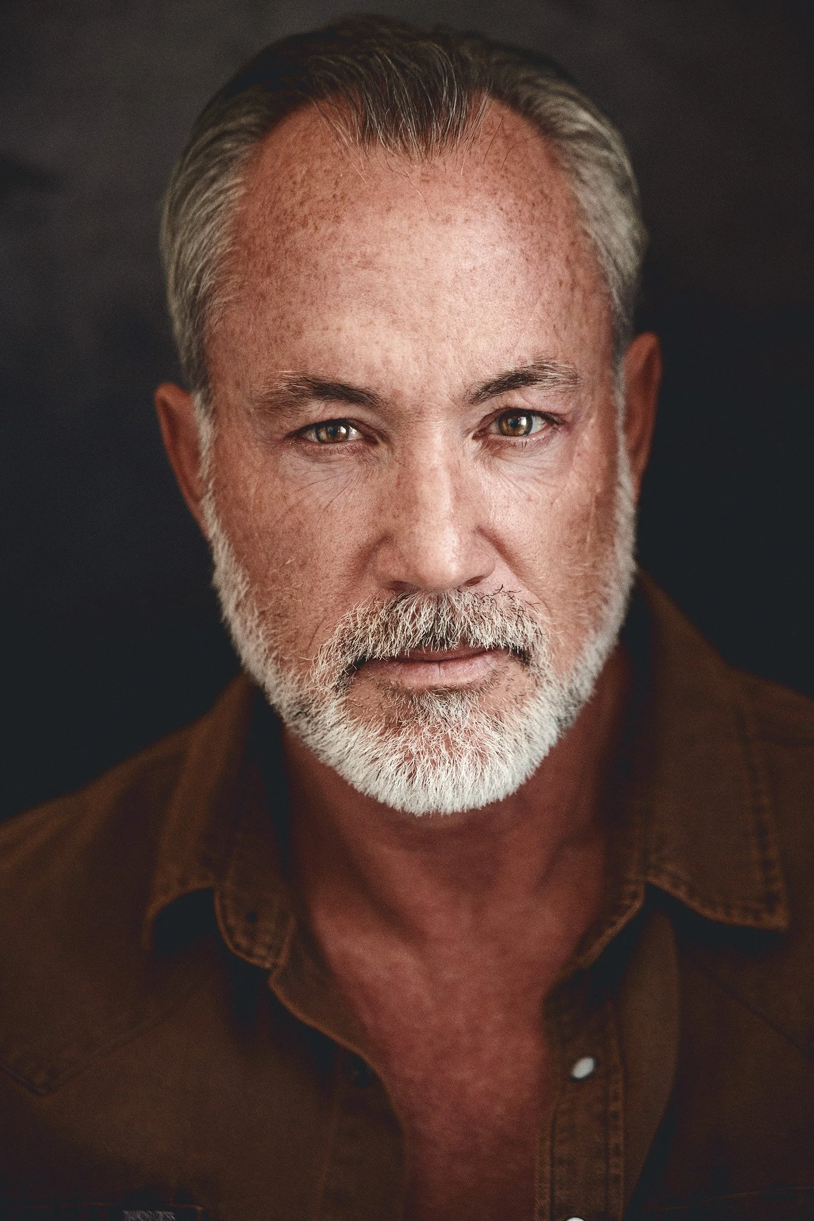 Close-up of a man with gray hair, gray beard, and mustache, wearing a brown shirt, looking directly at the camera. Dark background.