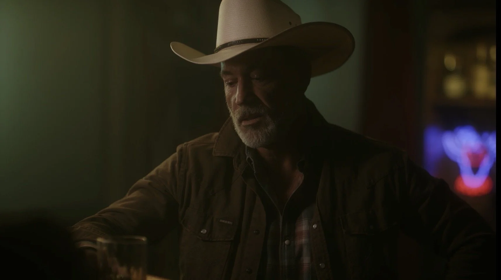 A man with gray beard wearing a cowboy hat and a brown jacket, sitting in a dimly lit bar or pub.