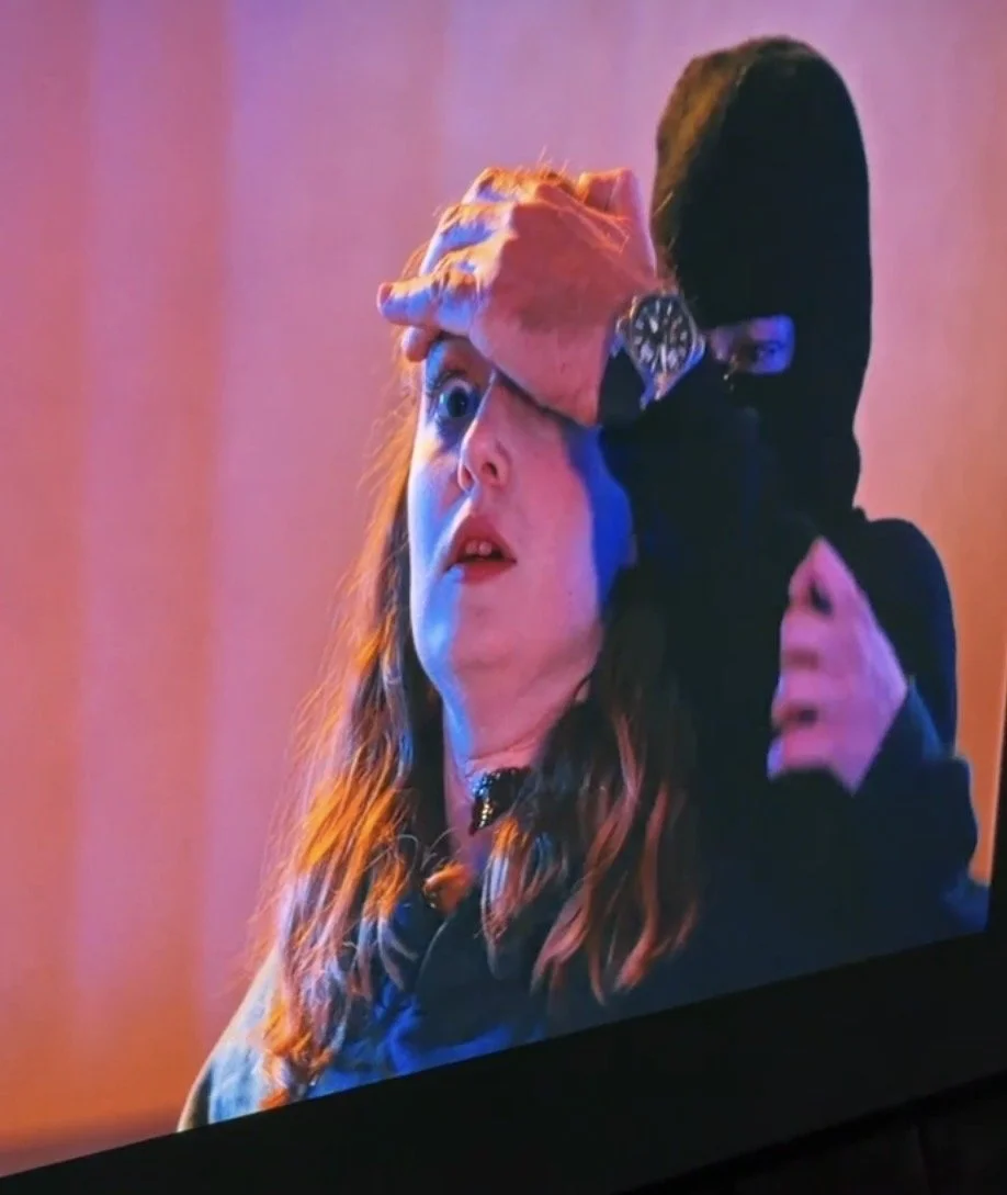 A woman with red hair and a choker necklace looks surprised, with her mouth slightly open. A person in a black hoodie, wearing a watch, covers her forehead with their hand. The background has pink and purple lighting.