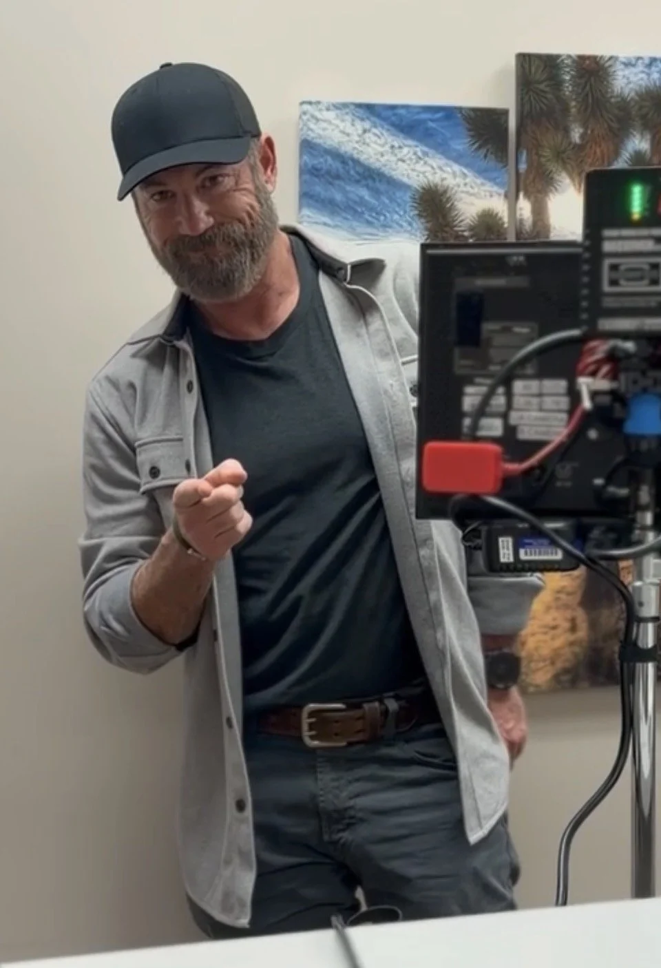 A man with a beard and a black cap smiling and pointing at the camera, standing in front of a camera setup with art of the desert and Joshua trees in the background.
