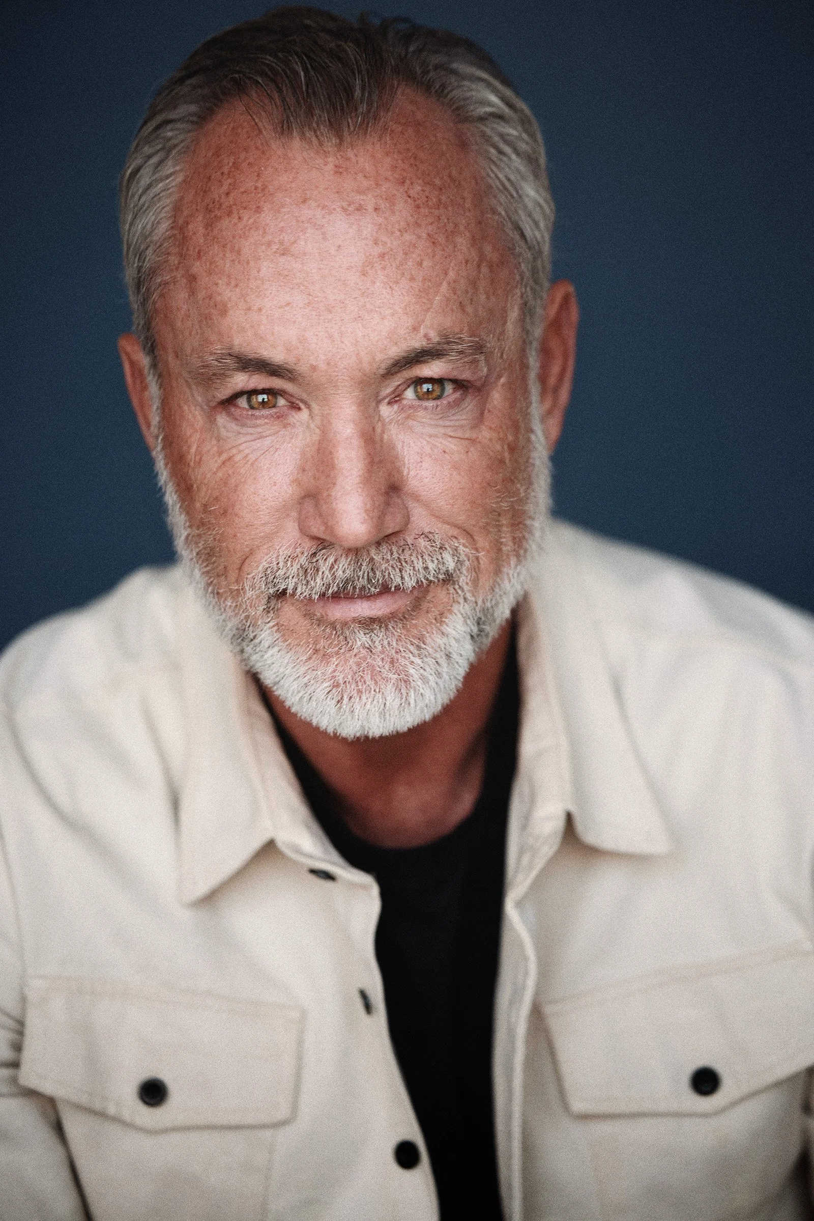 Close-up portrait of a man with gray hair and beard, wearing a white jacket over a black shirt, smiling softly against a dark background.