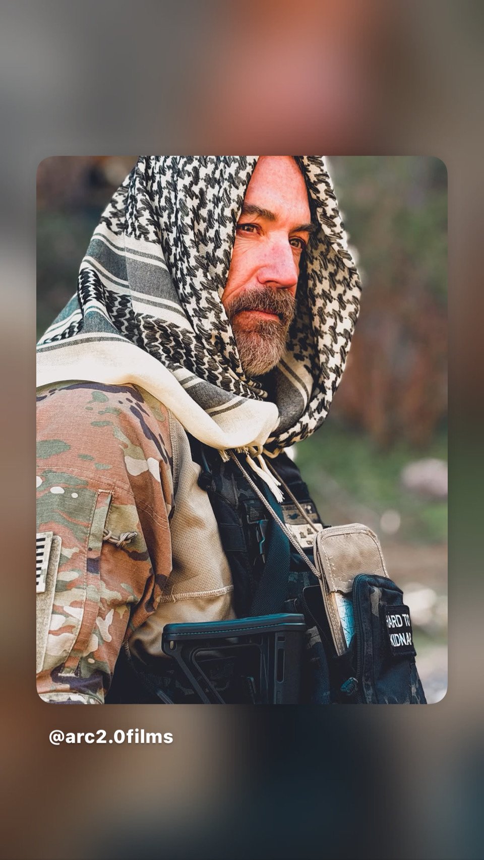 A man with a gray beard and mustache, wearing a black and white keffiyeh scarf and camouflage clothing, outdoors with a blurred natural background.