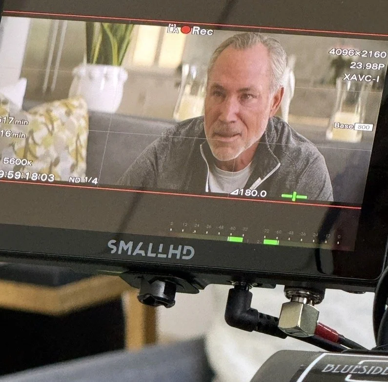 A man being filmed on a camera monitor during an interview or video shoot. The monitor displays technical information about the recording setup.