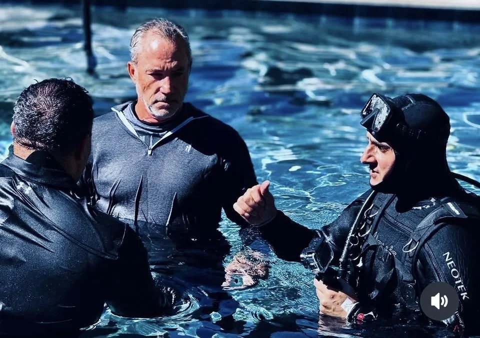 Three men in wetsuits in a swimming pool, one in the center with a serious expression, two others engaging with each other; one wearing a headlamp.