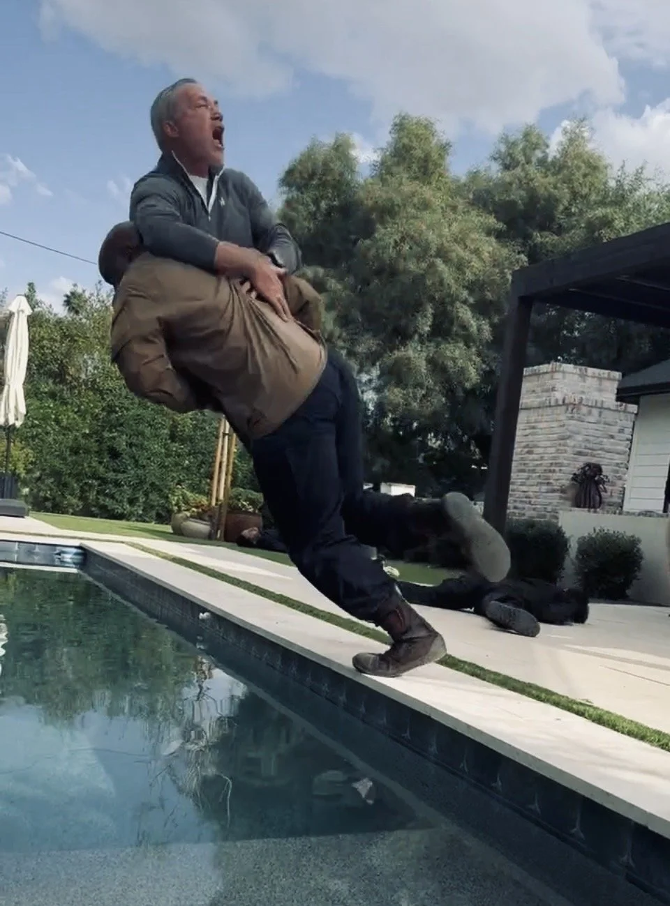 A man is being pulled into a swimming pool while another person is holding him around the waist. The man being pulled is screaming or yelling, and the scene appears to be humorous or staged.