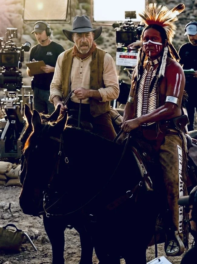 A scene on a film set with a man in cowboy attire and a Native American man with traditional face paint and clothing, both riding horses, surrounded by crew members and filming equipment.