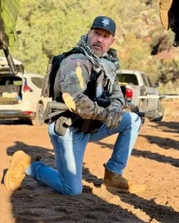 Man kneeling outdoors in casual clothes with a backpack, in front of parked cars and trees during daytime.