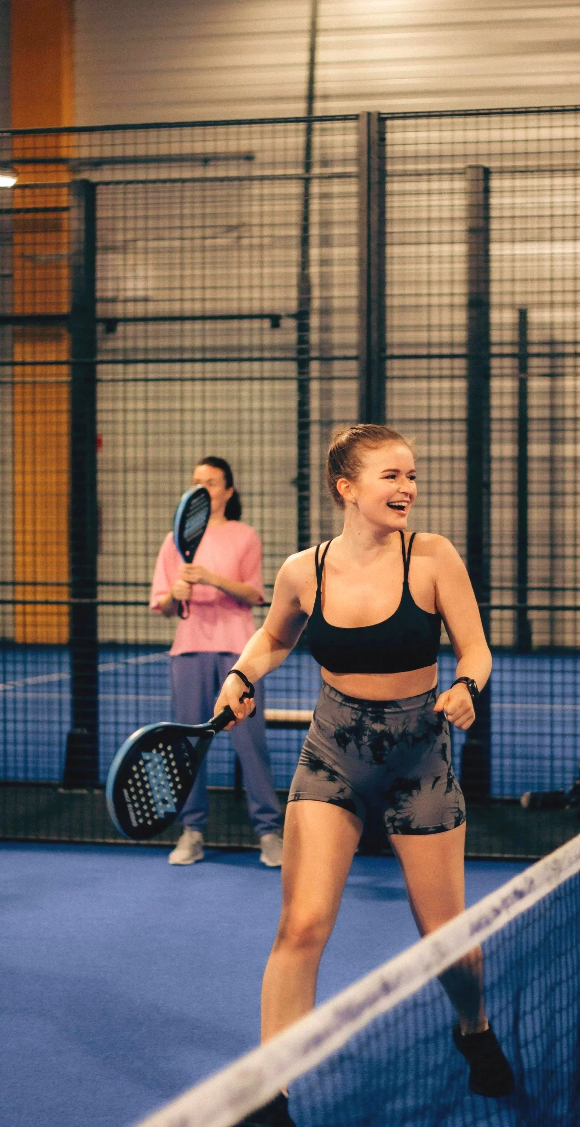 Deux femmes jouent au tennis en intérieur, une en avant et une en arrière. La femme en avant sourit, porte un haut noir et un short gris. La femme en arrière porte un haut rose et des pantalons gris, elle tient une raquette.
