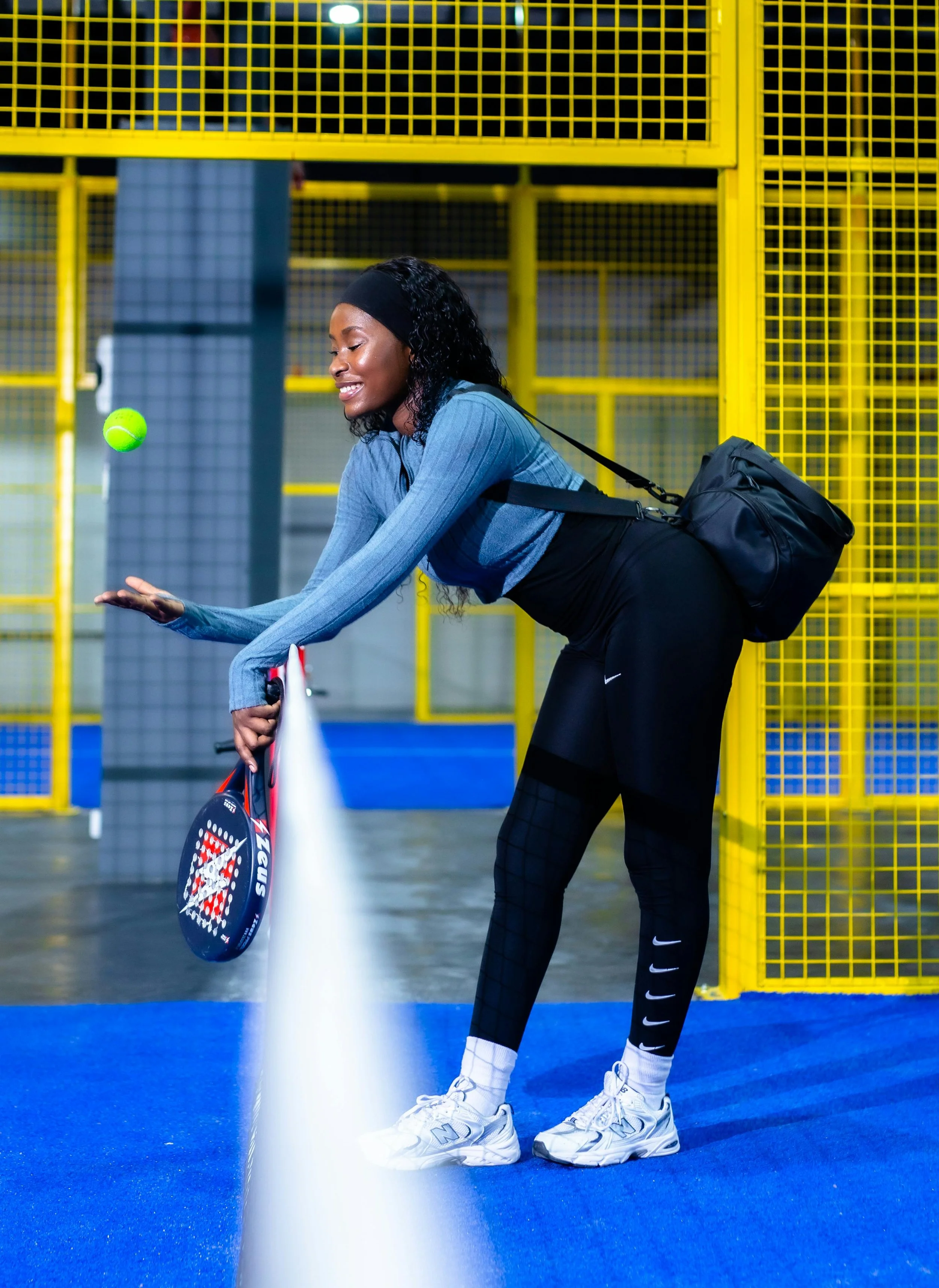 Une femme souriante en tenue de sport, portant des chaussures blanches et un sac à dos, joue au tennis sur un court intérieur avec un filet bleu, tandis qu'un ballon de tennis passe devant elle.