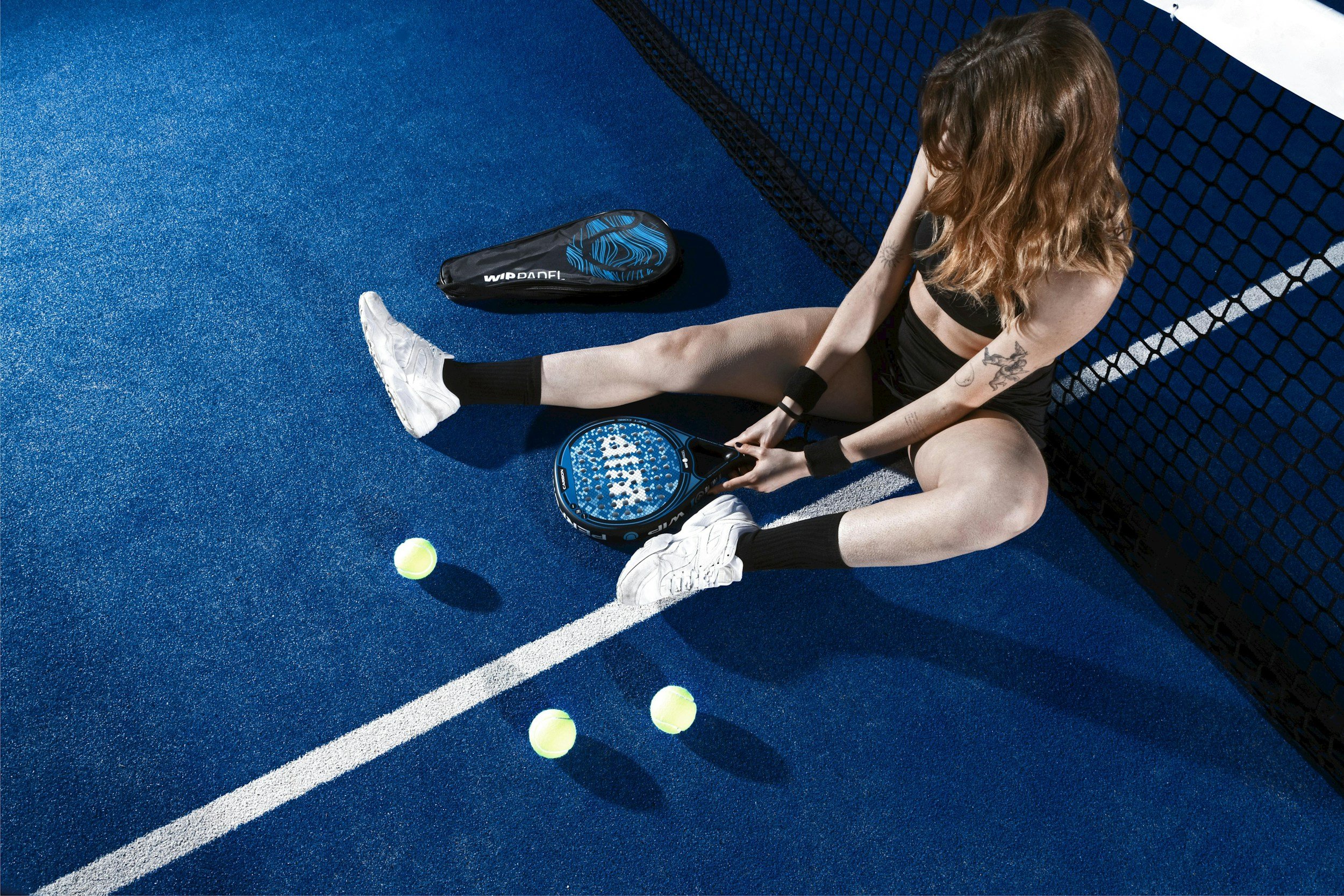 Une femme assise sur un terrain de tennis bleu, tenant une raquette, entourée de balles de tennis, portant une tenue sportive noire, des chaussures blanches, et ayant des tatouages sur le bras.