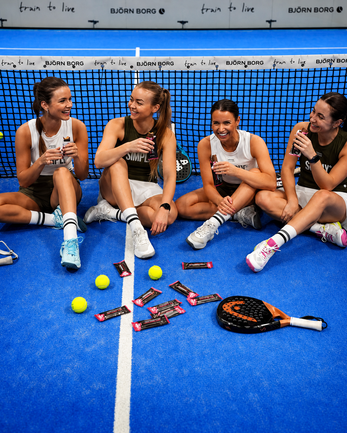 Quatre femmes assises sur un court de tennis en intérieur, souriantes et portant des tenues de sport, après une partie de tennis. Devant elles, sur le sol, il y a des balles de tennis, des barres énergétiques, des raquettes et des bouteilles de boisson énergisante.
