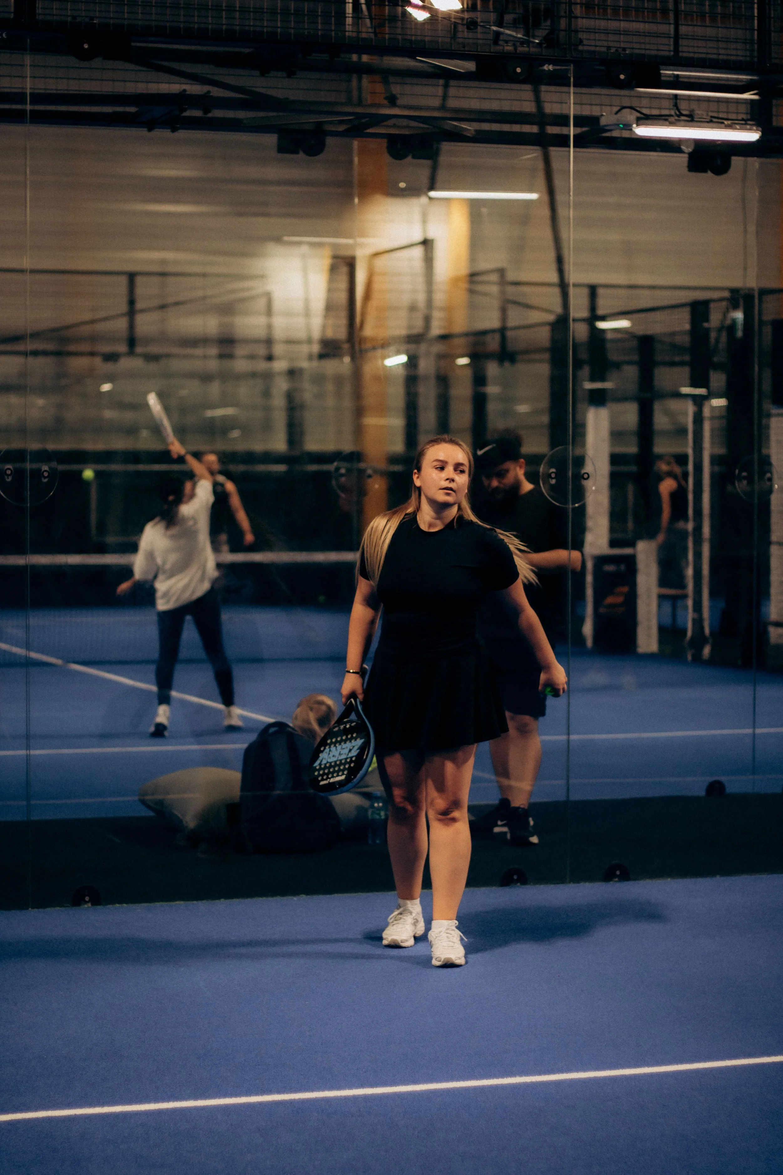 Jeune femme blonde portant un t-shirt noir, jupe noire et baskets blanches, debout dans une salle de tennis. On la voit à travers une vitre, avec des sacs et une raquette de tennis à ses pieds. En arrière-plan, d'autres personnes jouent au tennis dans une salle couverte.