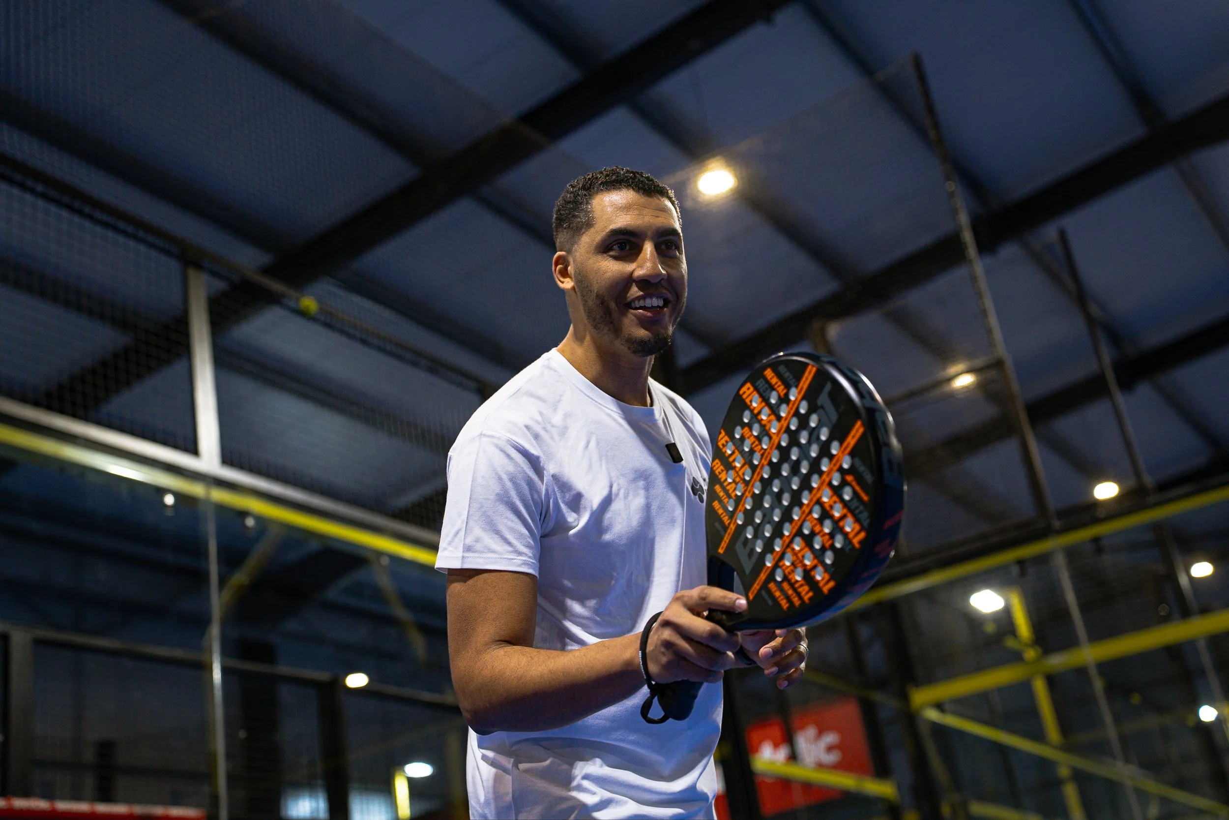 Un homme souriant tient une raquette de paddle tennis dans une installation sportive intérieure.