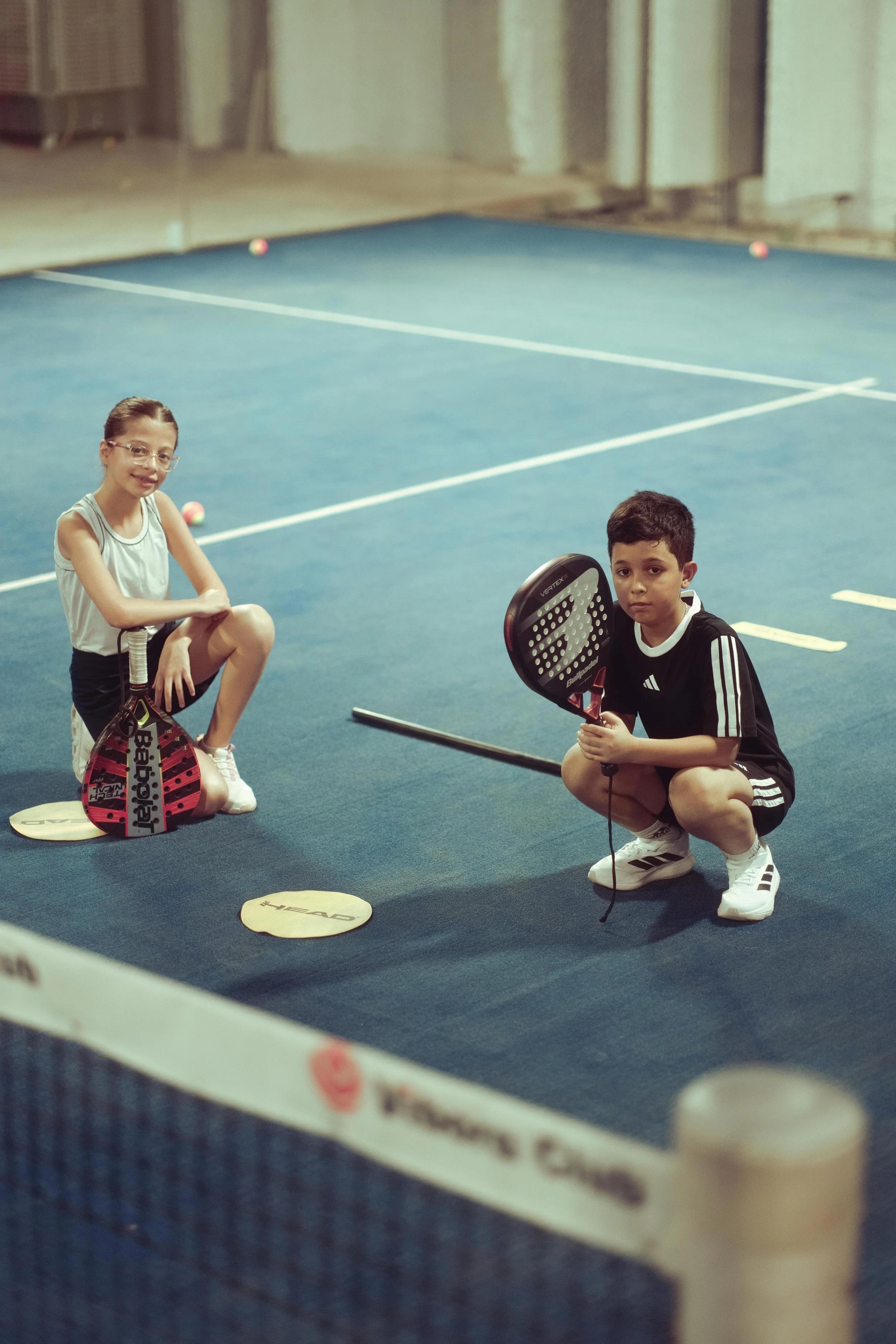 Deux enfants, un garçon et une fille, posent avec des raquettes de tennis sur un court intérieur, environnés de lignes de jeu et de balles de tennis.