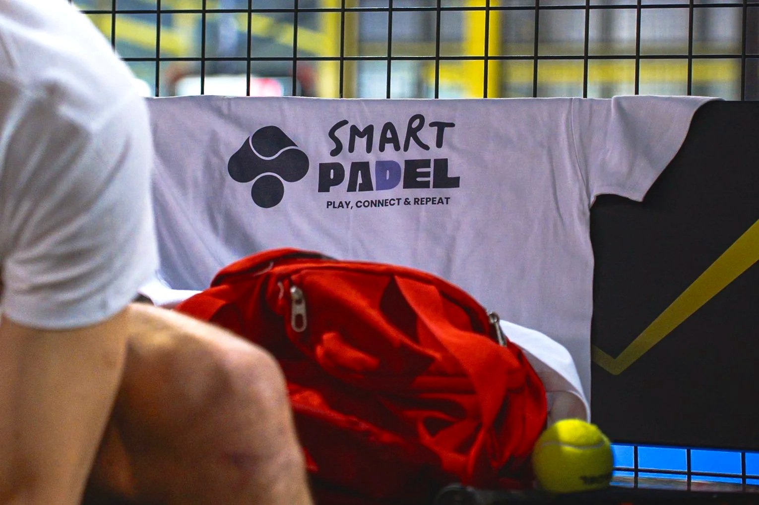 Une table avec un t-shirt blanc portant le logo et le nom de 'Smart Padel' et le slogan 'Play, Connect & Repeat'. En premier plan, un sac de sport rouge et une balle de padel jaune, avec un joueur en arrière-plan.