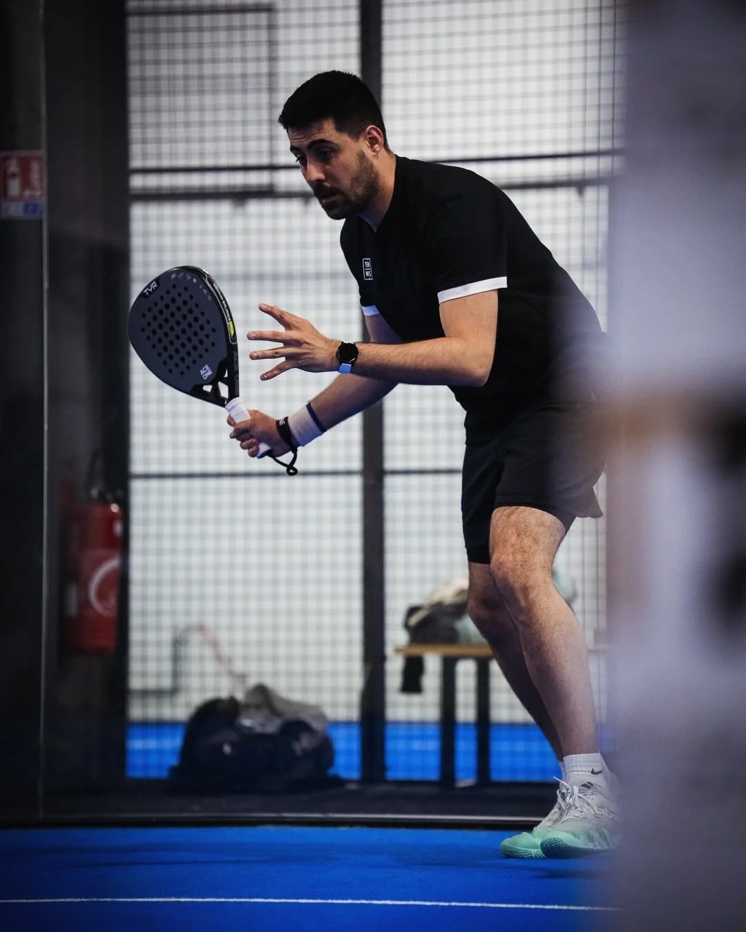 Homme jouant au paddle-tennis, en position prête à frapper la balle, portant une tenue de sport noire, chaussures blanches à semelles vertes, dans une salle d'entraînement.