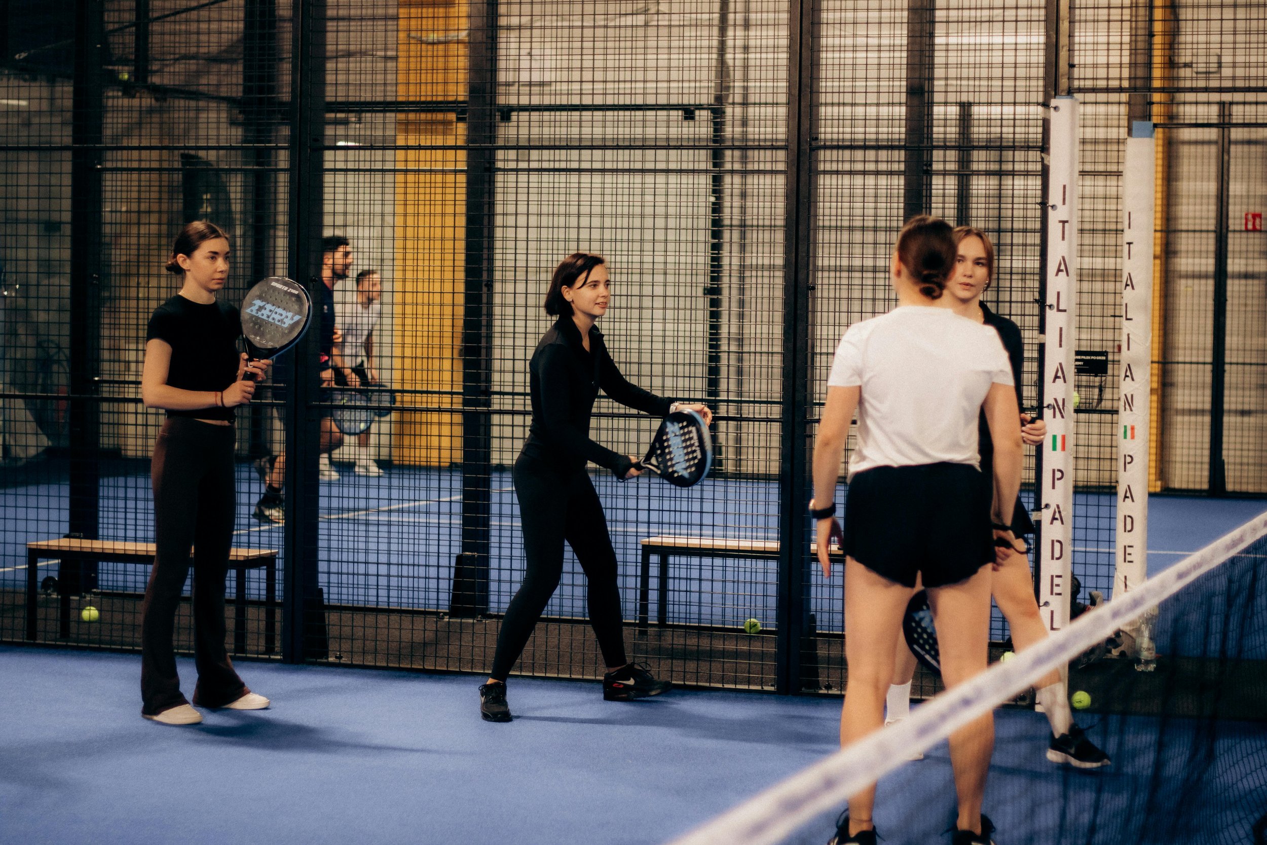 Des jeunes femmes discutent près d'une barrière dans un terrain de padel indoor, équipées de raquettes. Elles portent des vêtements de sport, et il y a des balles de tennis au sol.