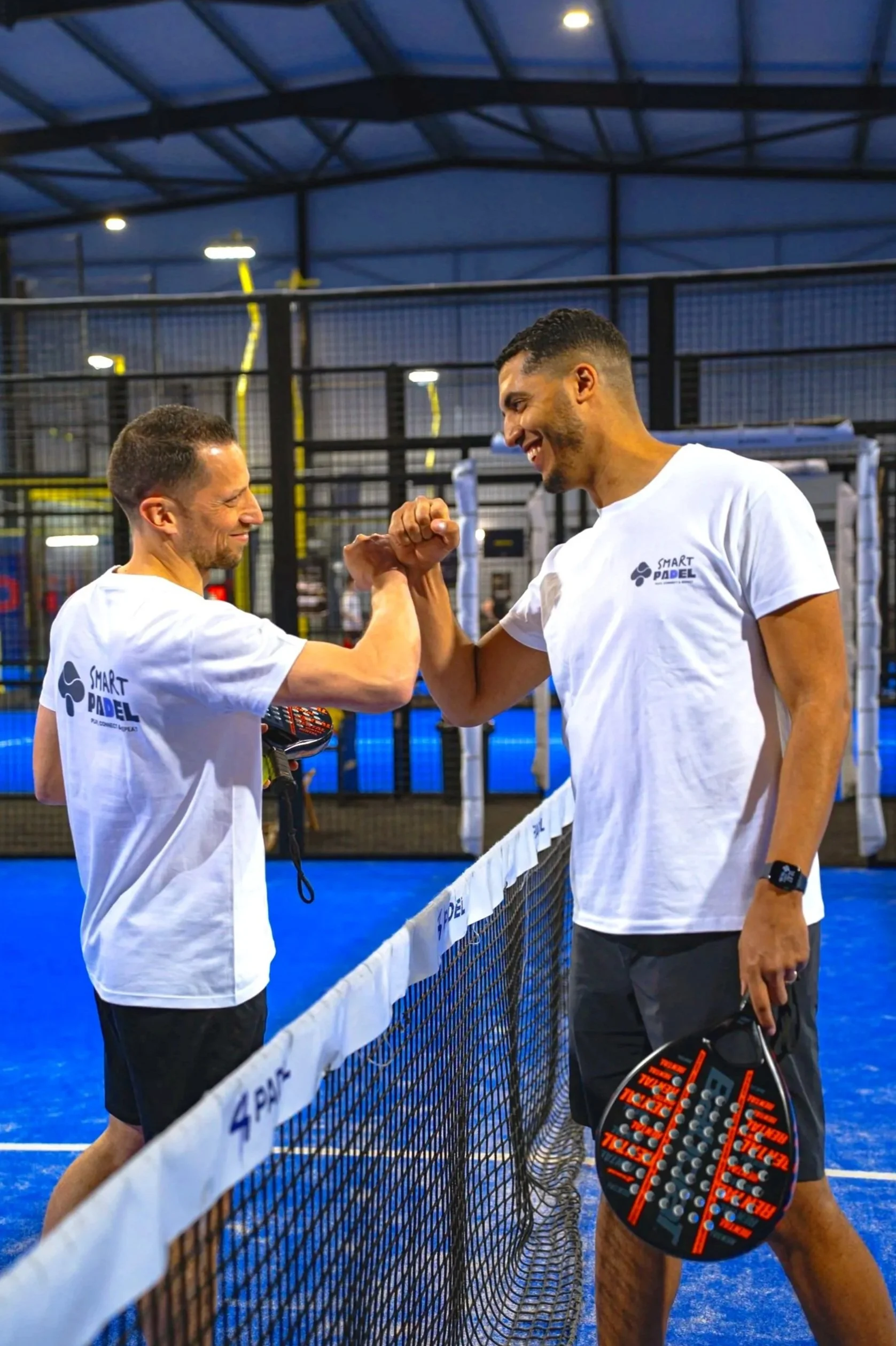 Deux hommes souriants se saluant et se donnant un coup de poing dans un centre de padel, portant des t-shirts blancs avec le logo Smart Padel, un filet de padel en premier plan et une structure en métal en arrière-plan.
