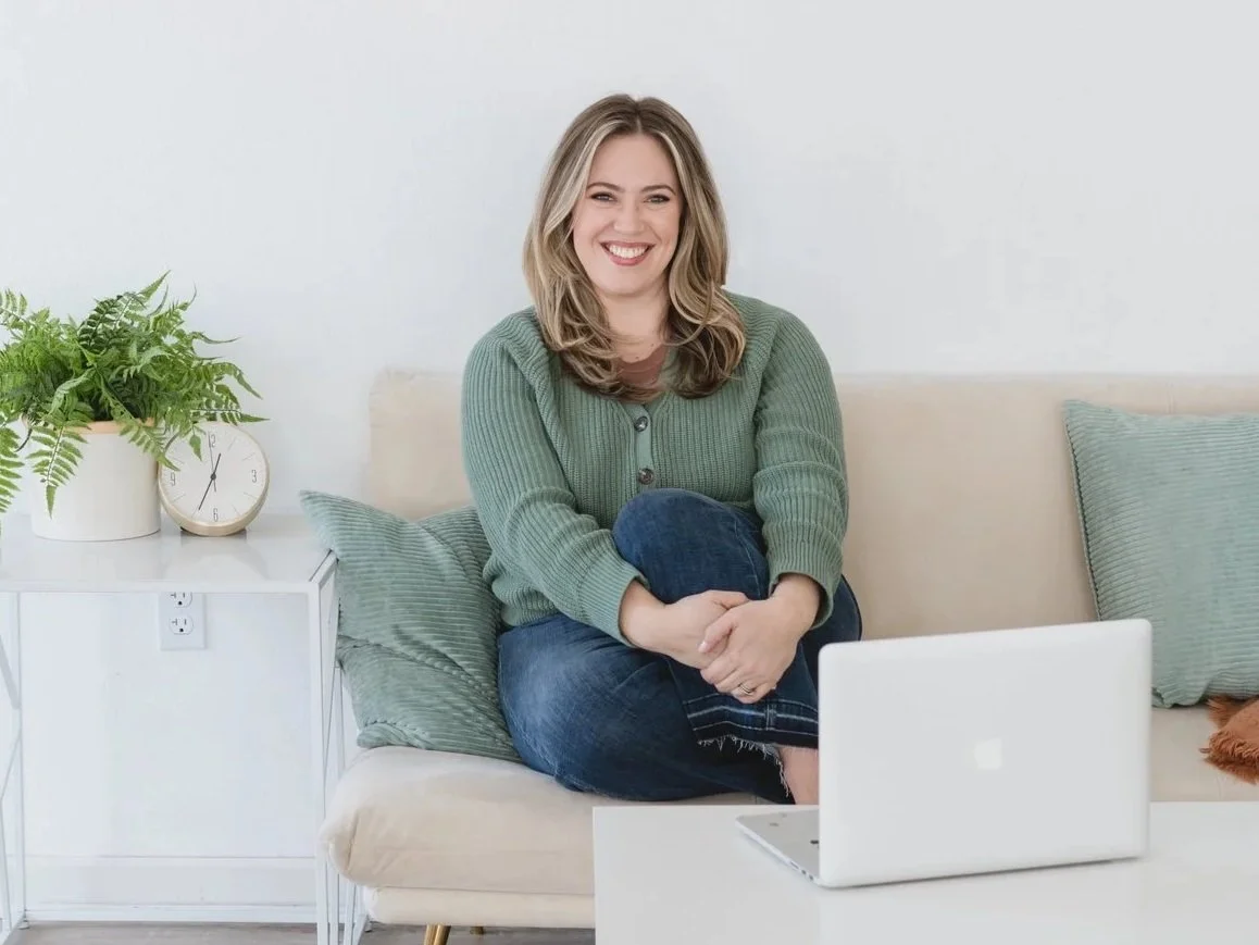 A woman sitting on a beige sofa with cushions, smiling, with a green sweater, in a modern living room. There is a white side table with a green potted fern, a clock, and a plugged-in white laptop in front of her.