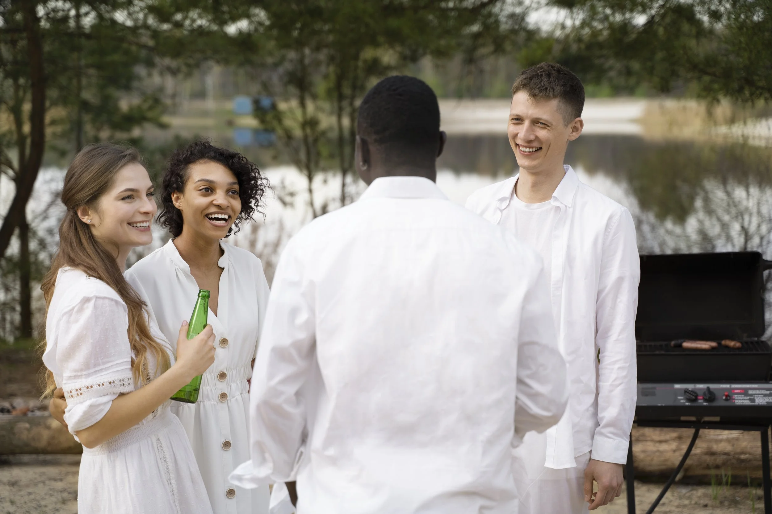 Group of people at an outdoor gathering, smiling and talking, with a grill and trees in the background.