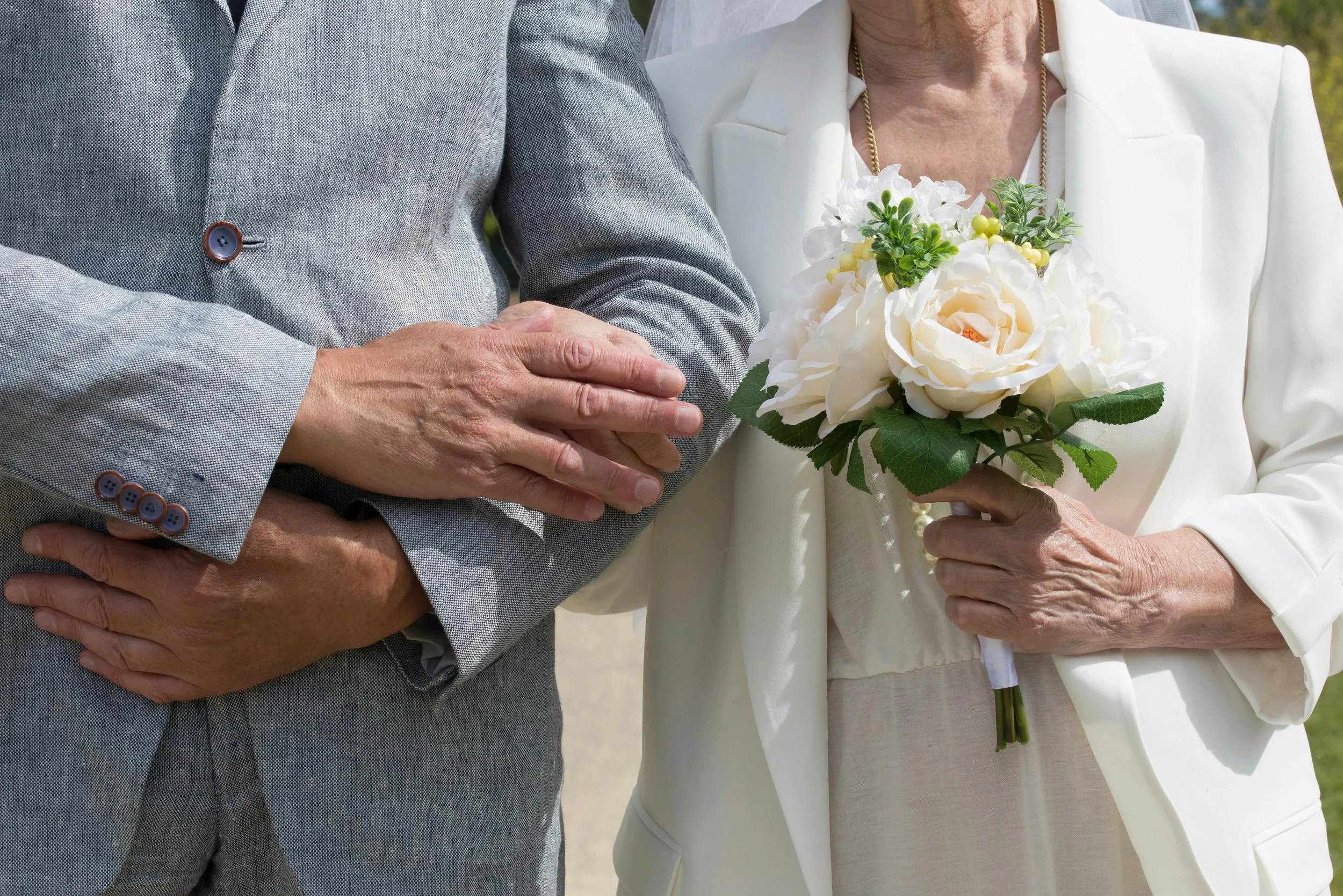 Close-up of a man and a woman holding hands, with the woman holding a bouquet of white flowers, outdoors.