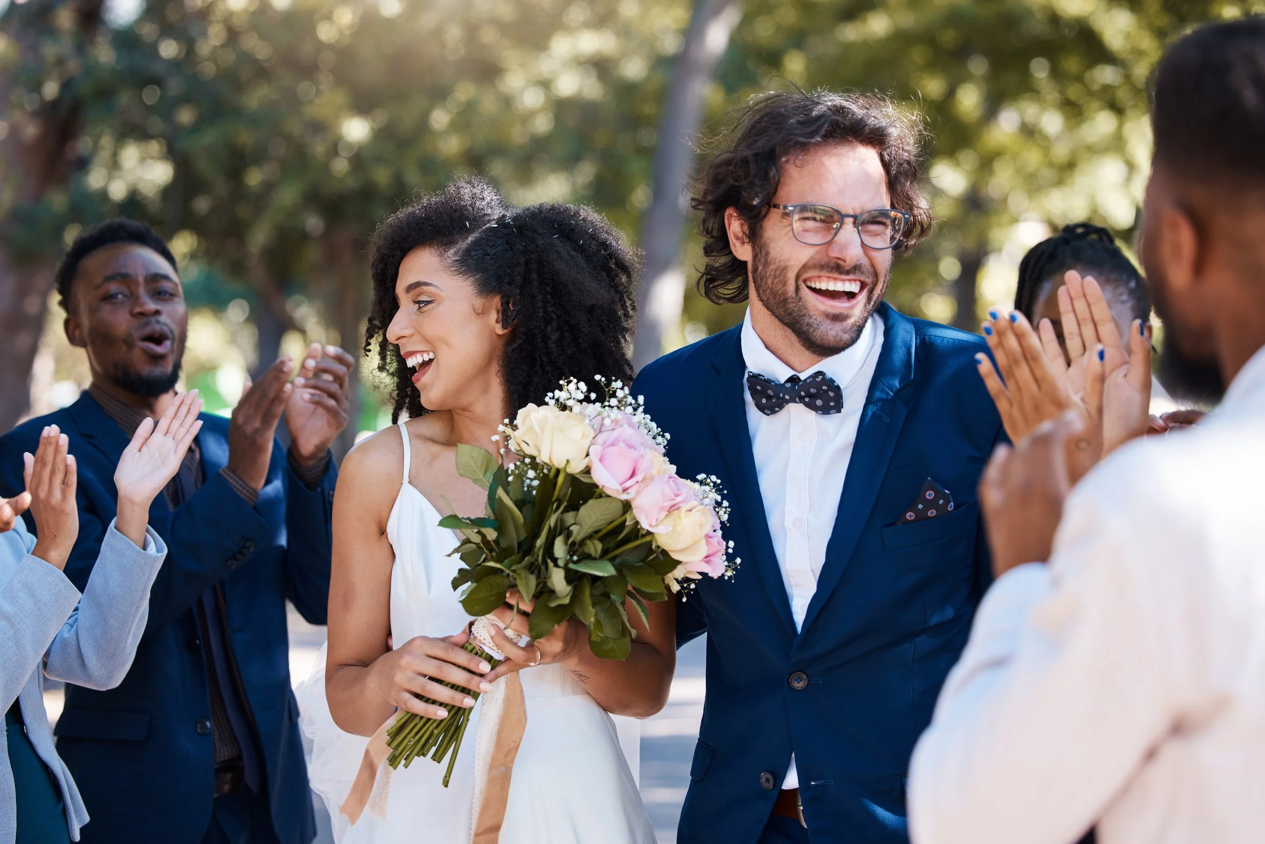 Bride holding a bouquet of flowers, surrounded by people clapping and celebrating outdoors in a park.