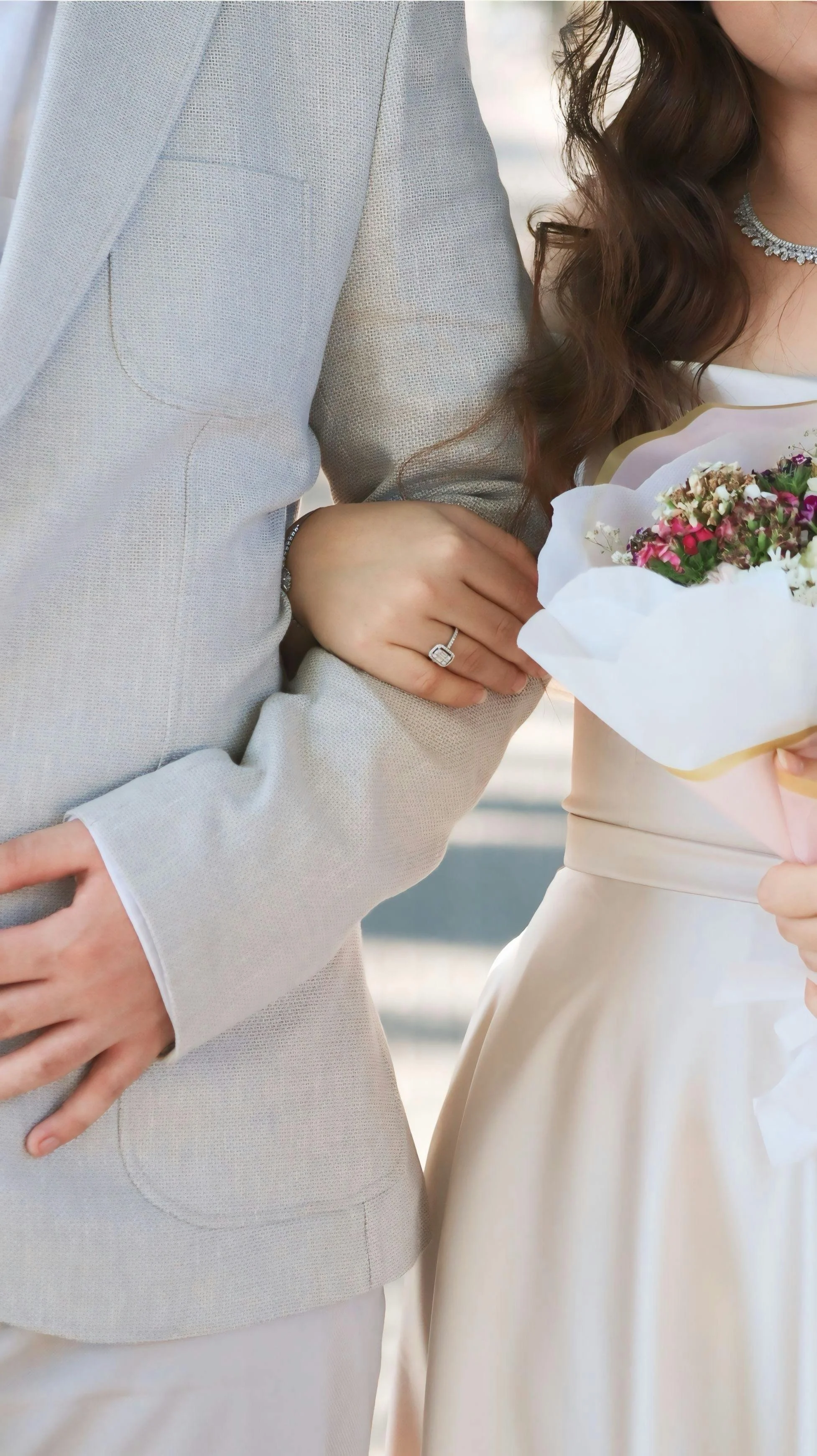 Close-up of a couple holding hands, a woman with a diamond ring, a man in a light-colored suit, woman holding a bouquet of flowers, rear view.