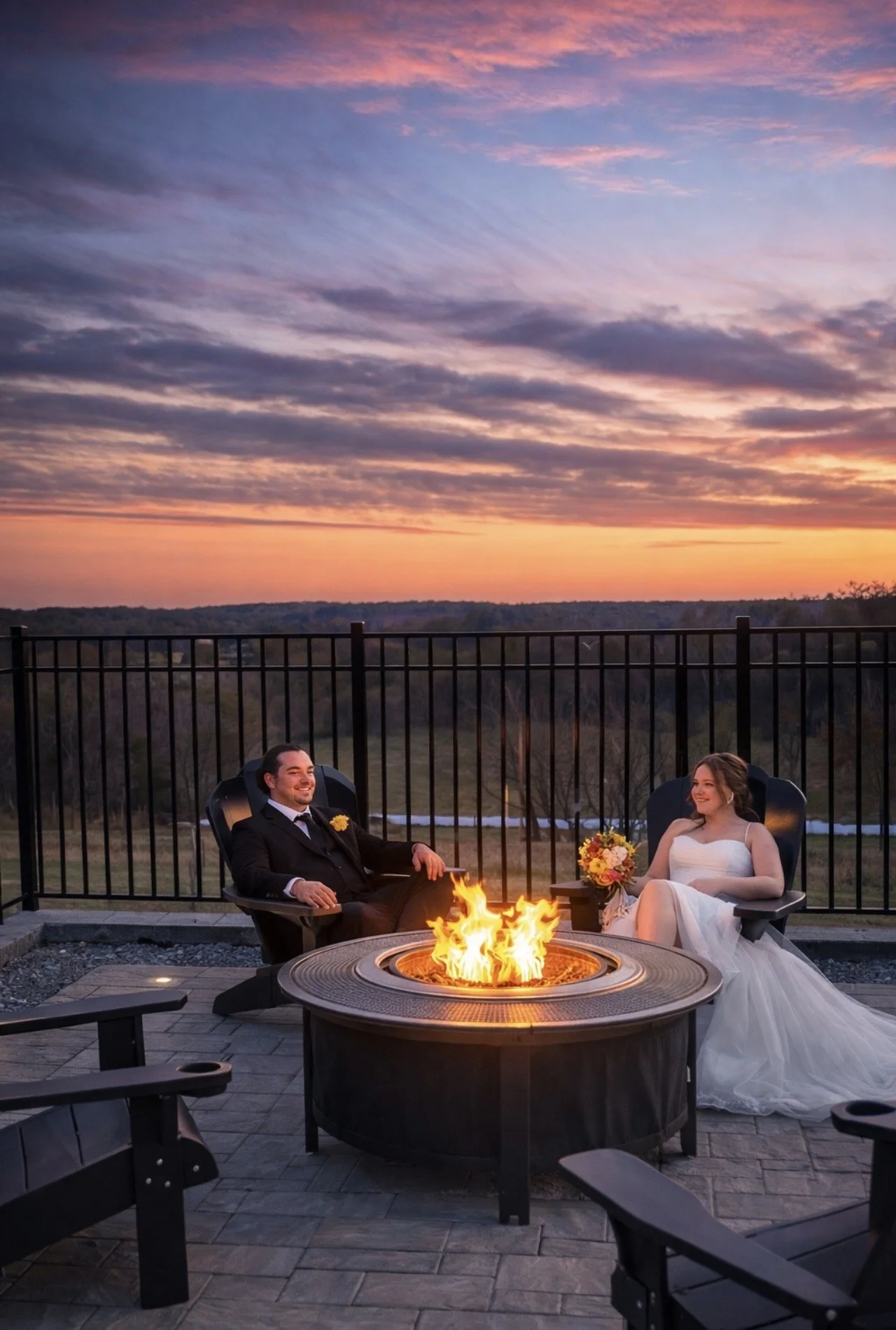 A bride and groom sitting near a fire pit on an outdoor patio during sunset. The groom is in a black suit and the bride in a white wedding dress, holding a bouquet. The sky is colorful with shades of pink, purple, and orange, and a black metal fence surrounds the patio.