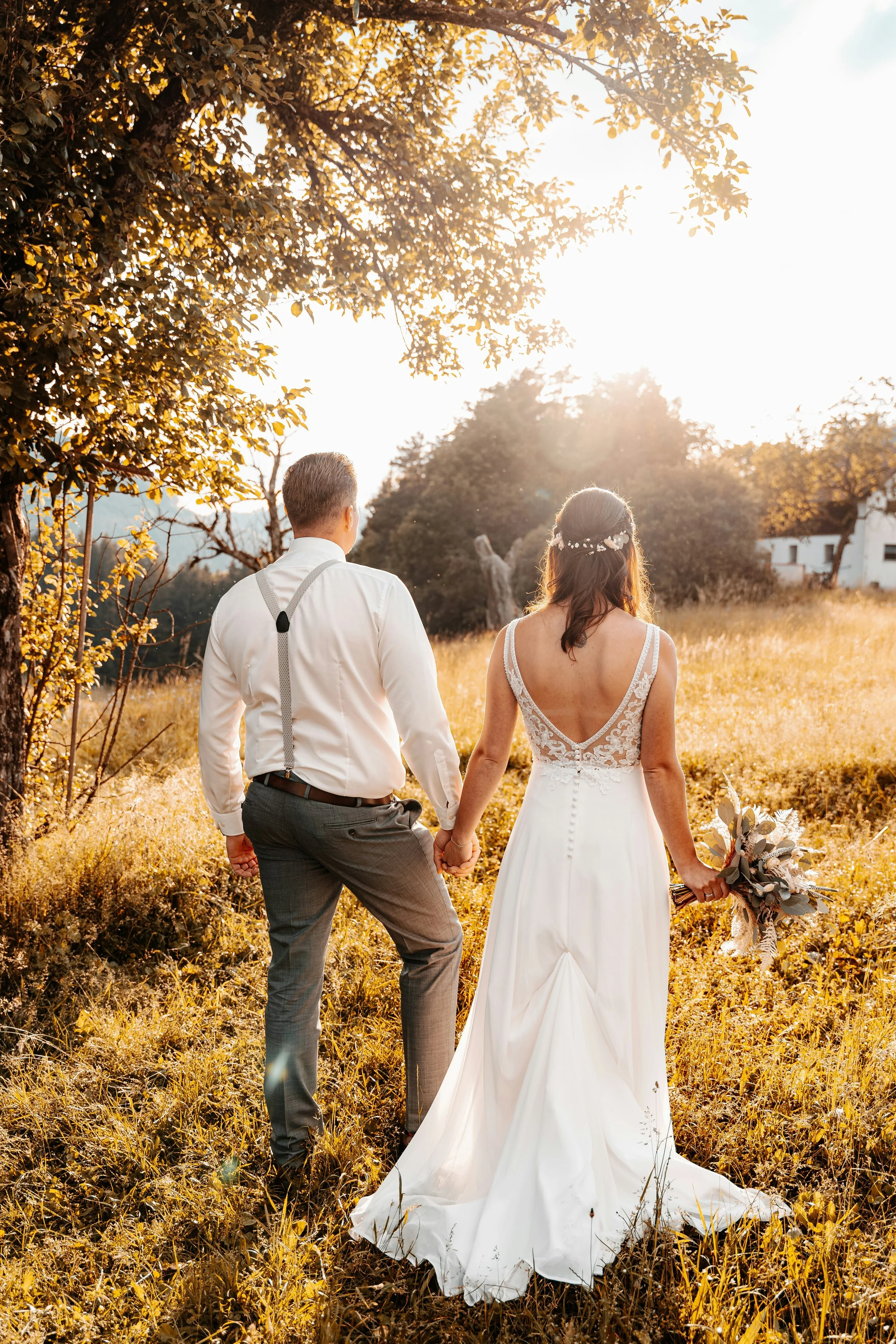 A bride and groom holding hands and walking in a golden field during sunset, with trees and a white building in the background.