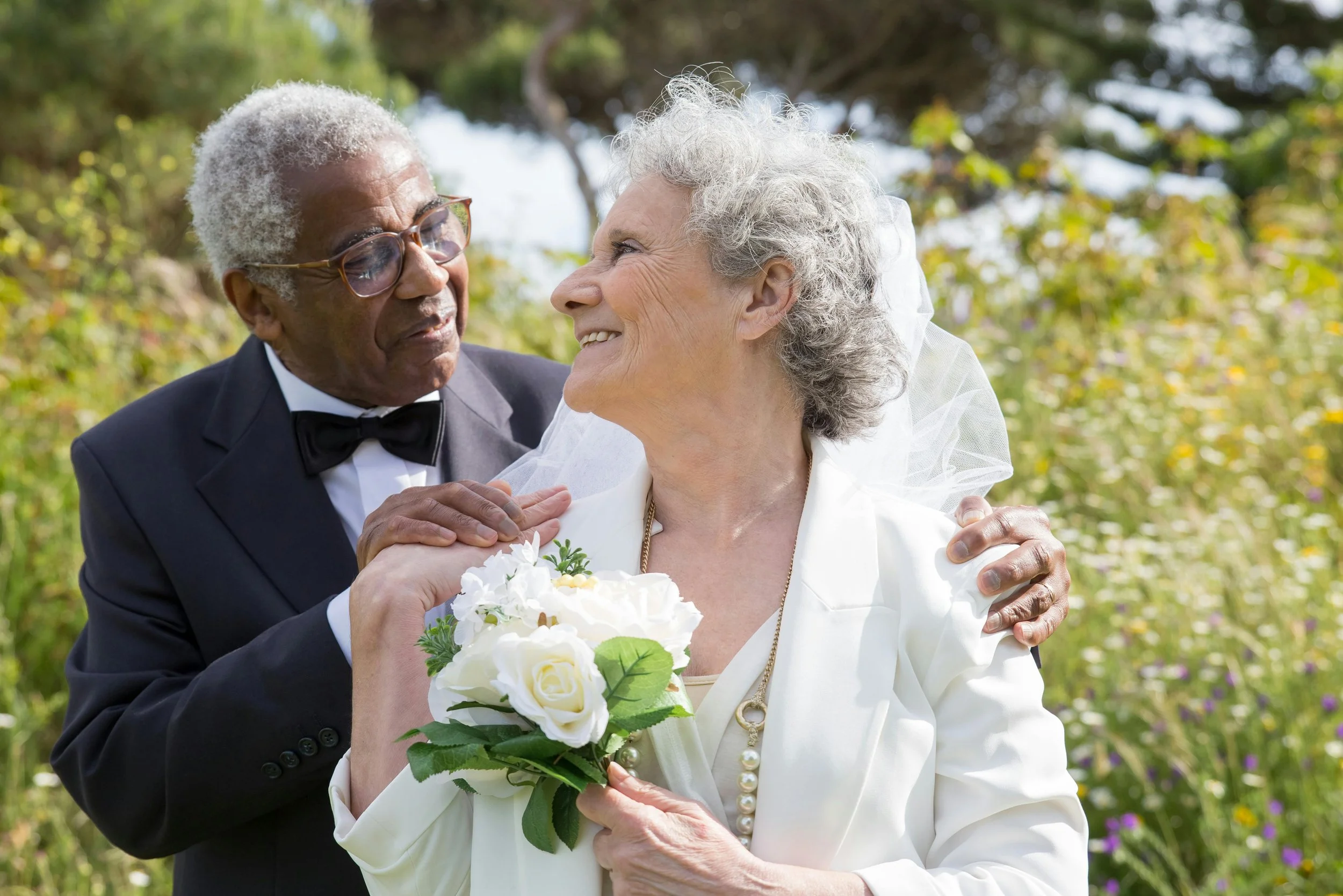 An elderly couple, a man in a tuxedo and a woman in a white dress holding a bouquet, sharing a joyful moment outdoors with trees and greenery in the background.