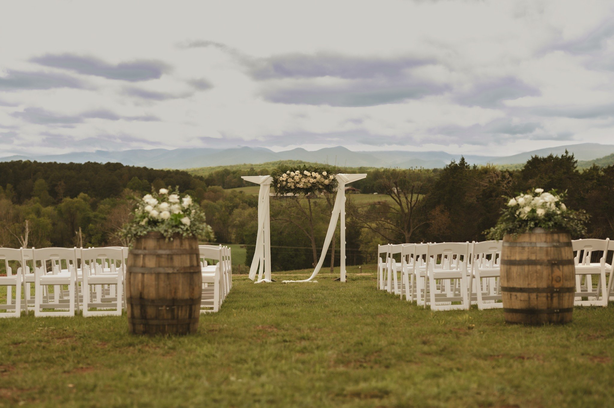 Wedding ceremony setup outdoors with white chairs, a floral arch, wine barrels, and a scenic mountain view in the background.