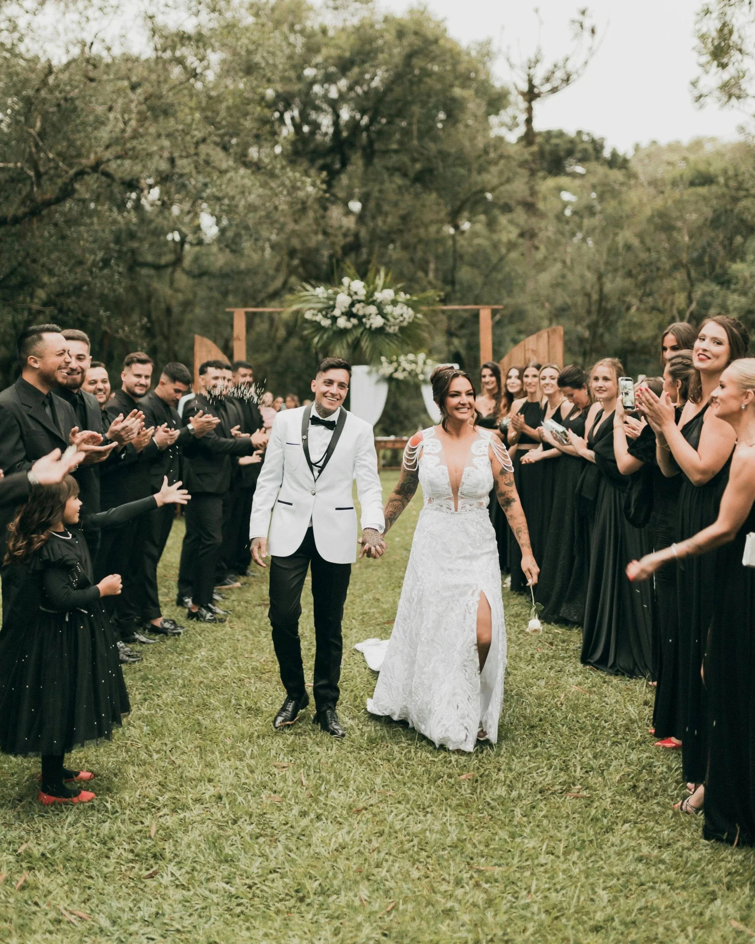 A wedding ceremony outdoors with a bride and groom walking hand-in-hand through a crowd of guests, surrounded by trees and greenery.