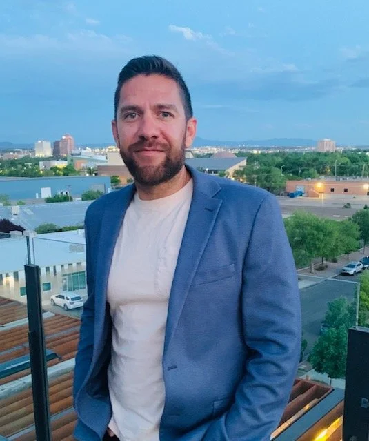 A man with dark hair and a beard standing outdoors on a balcony with a cityscape in the background, wearing a blue blazer and white shirt during the evening.