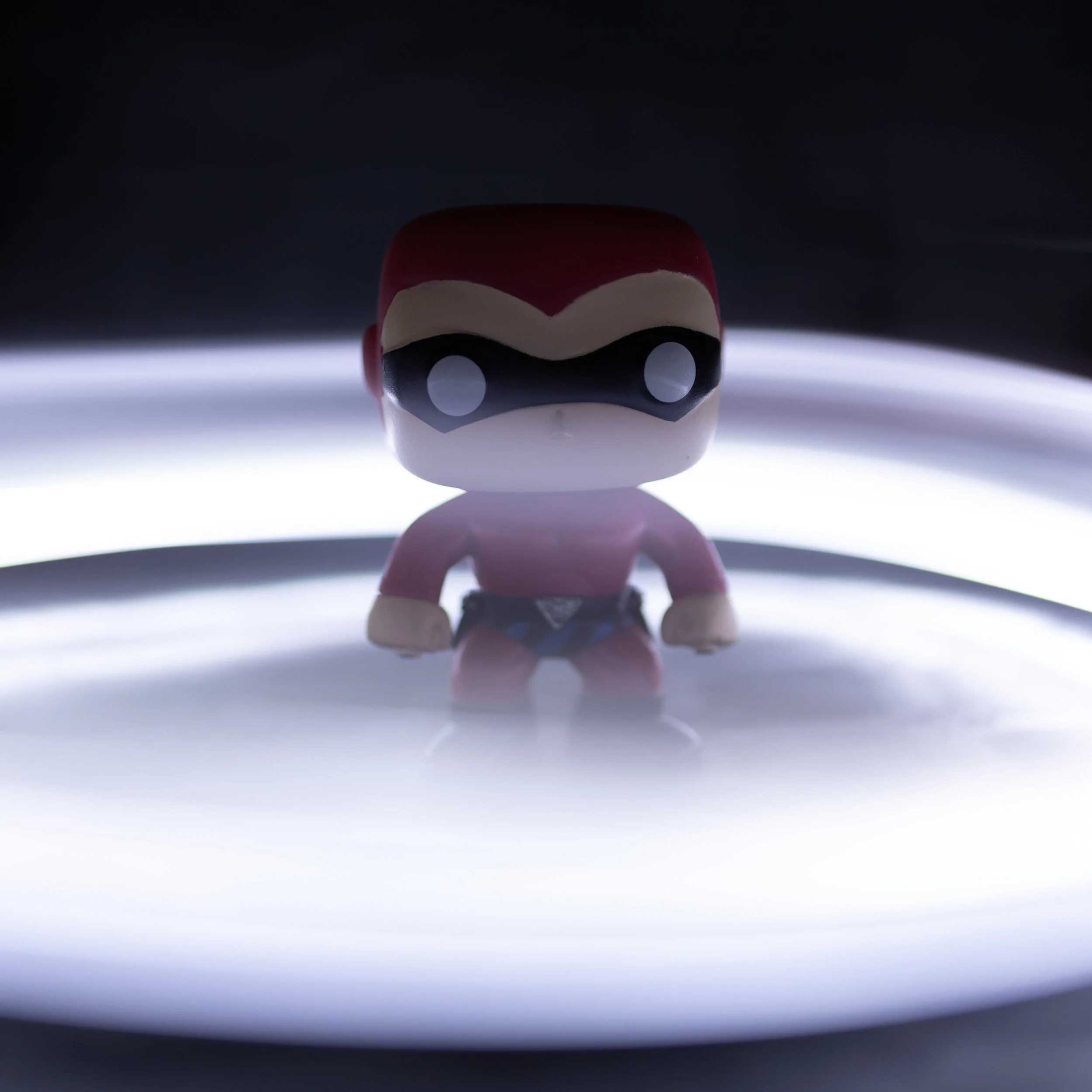 A small black, red, and white figurine with large white eyes, standing on a reflective surface with a bright light behind it, creating a high-contrast scene.