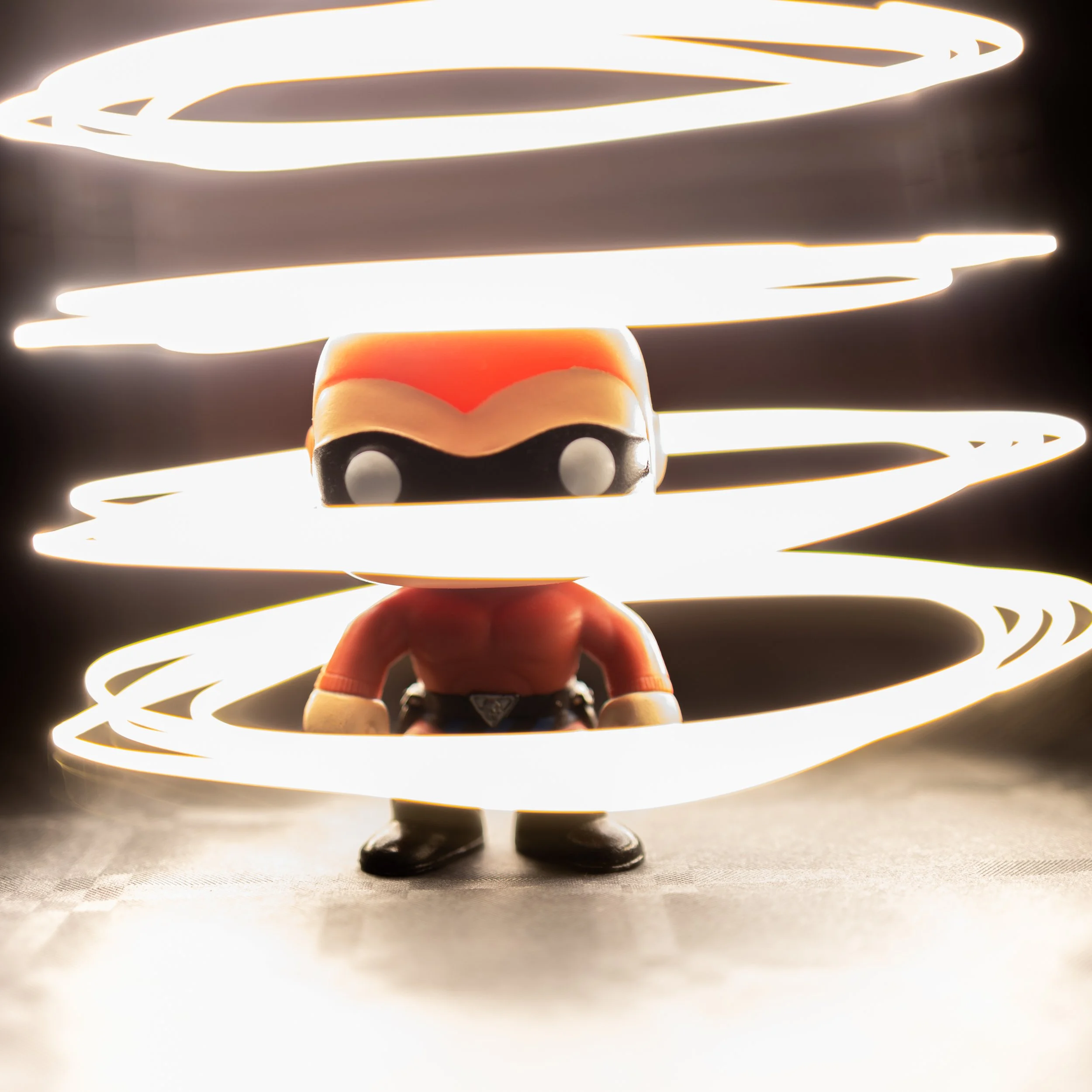 Close-up of a small action figure with a red chest, black eyes, black lower face, black shoes, and a cape, surrounded by glowing light streaks.