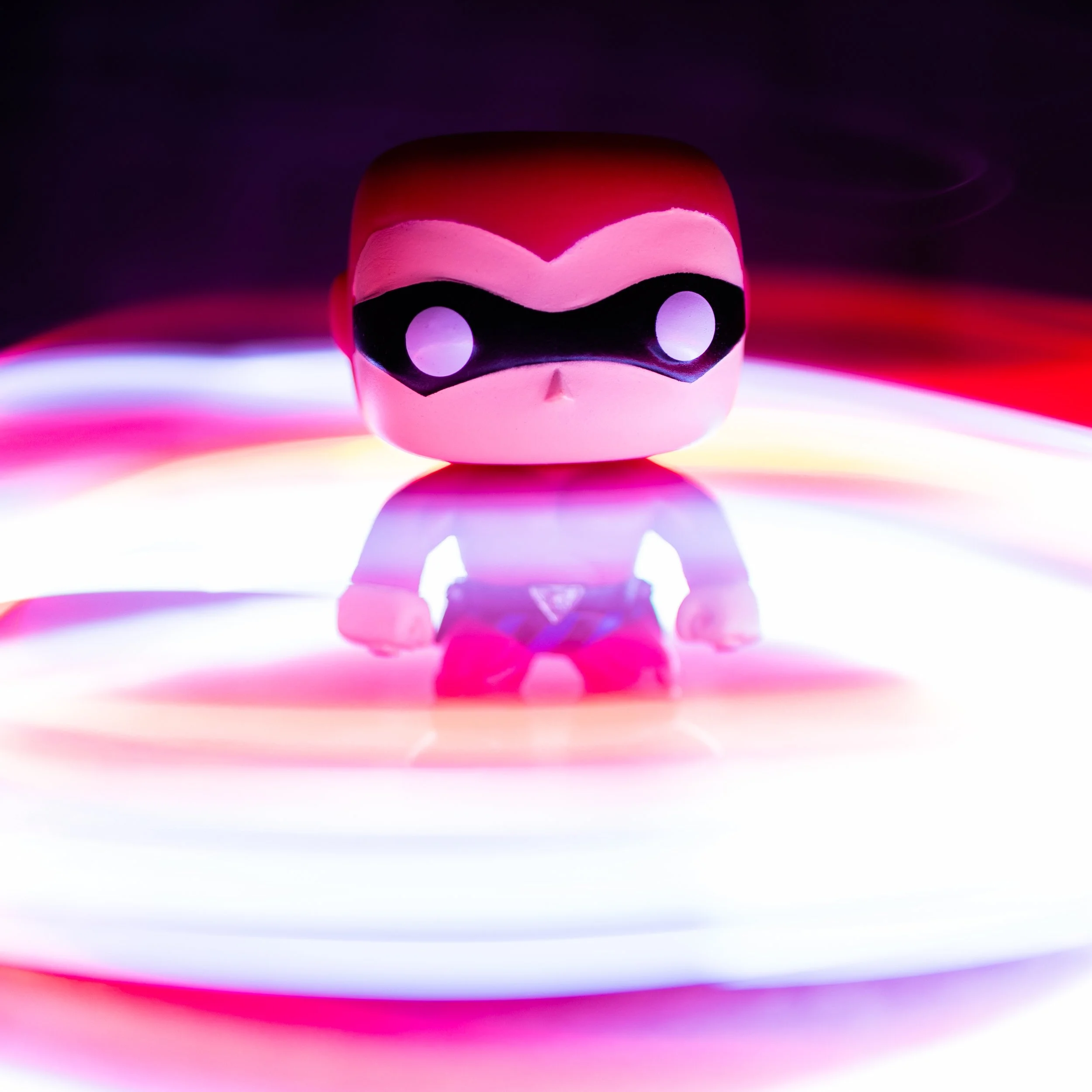 Close-up of a stylized, cartoonish pink and black skull figure with large, round eyes, set against a vibrant, multicolored background with swirling light effects.