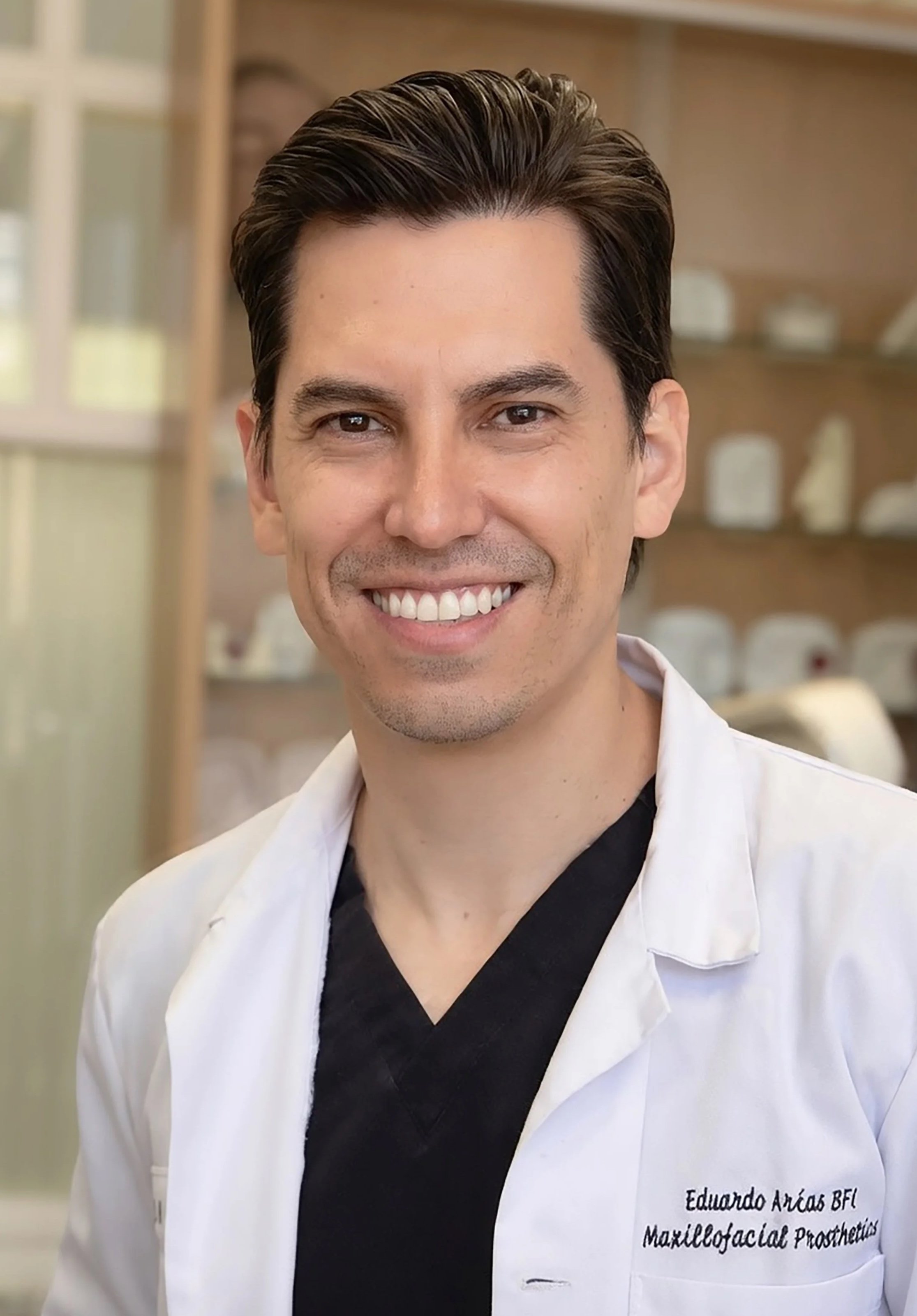 A man smiling, wearing a white medical coat with black text on it that says 'Eduardo Arias-Amezquita, CCA Maxillofacial Prosthetics'. Background appears to be a medical or clinical setting.