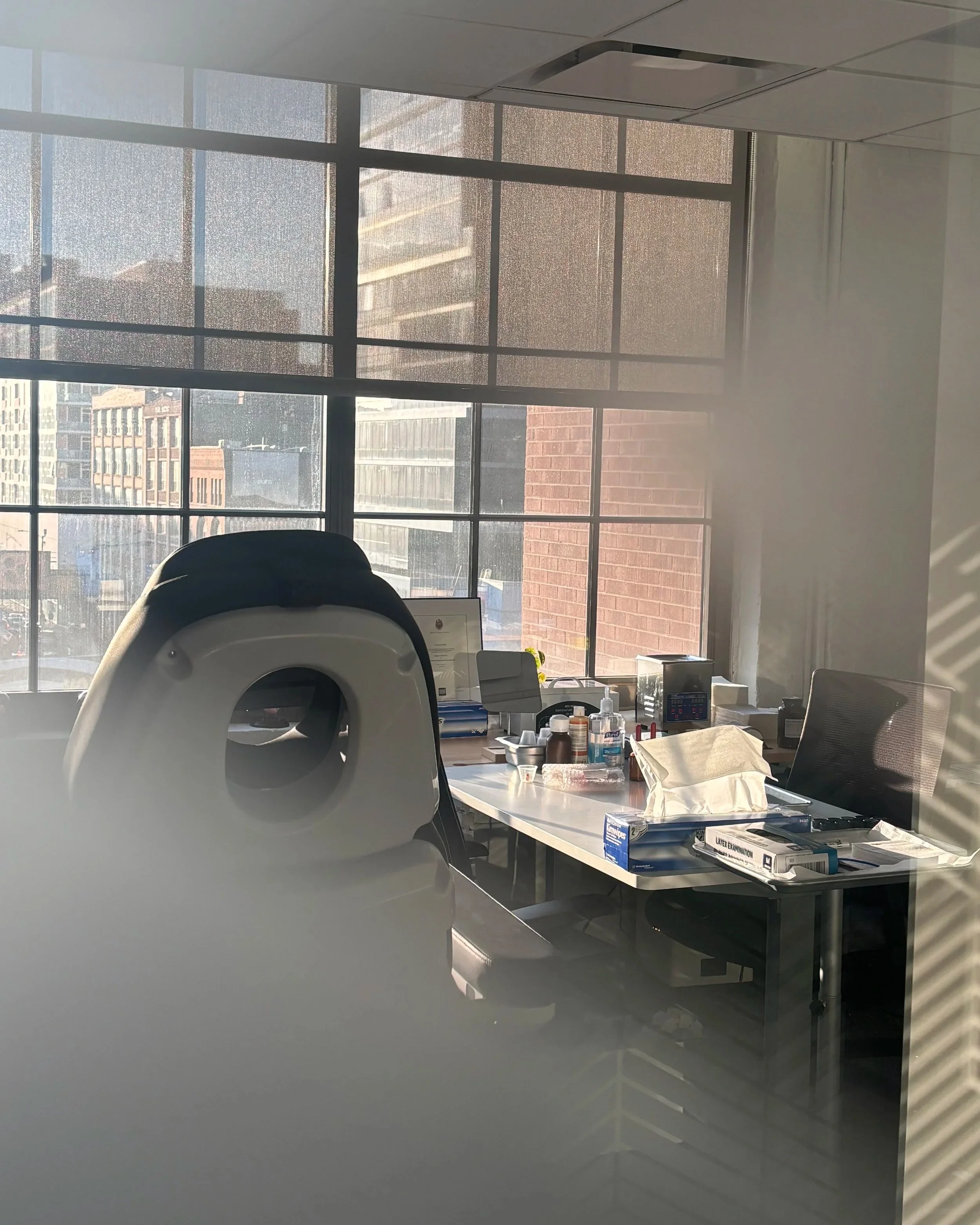 Office interior with a large window showing city buildings, a patient exam chair, a table with medical supplies, and office chairs.